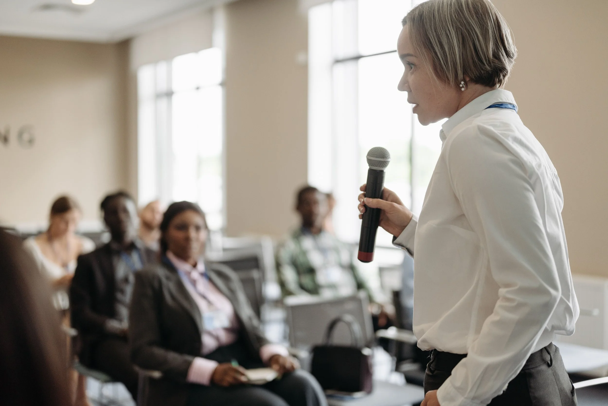 A woman in business attire is speaking into a microphone during a presentation or seminar in a well-lit room with large windows. Audience members are seated and listening.