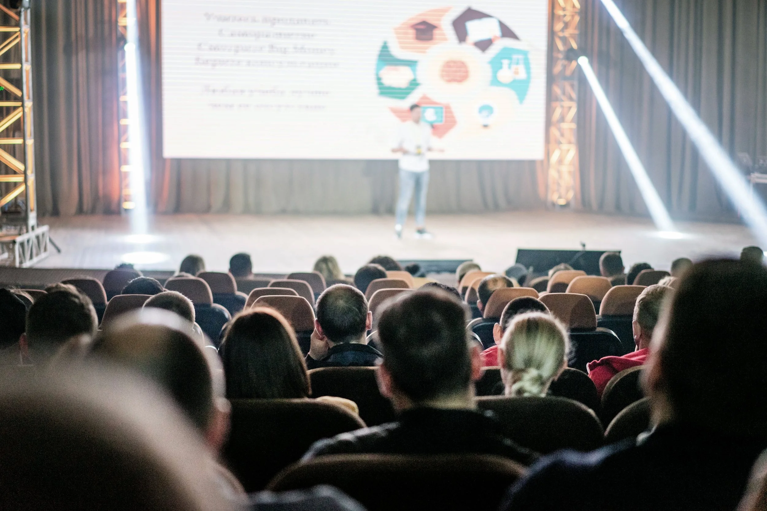 A man is giving a presentation on stage in front of an audience seated in an auditorium. The background features a large screen with text and colorful graphics.