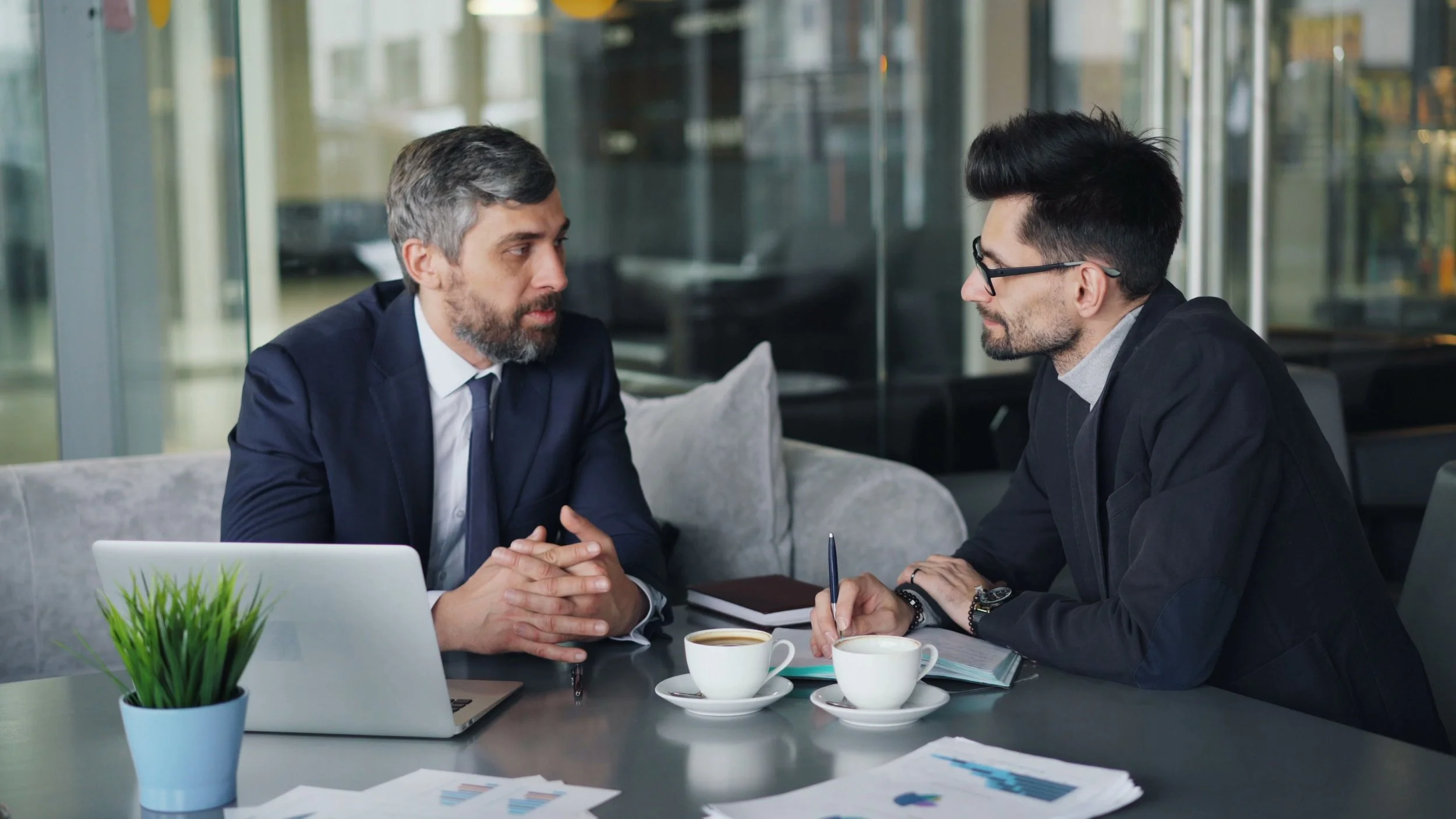 Two men having a serious business discussion in a modern office conference room, surrounded by documents and cups of coffee.