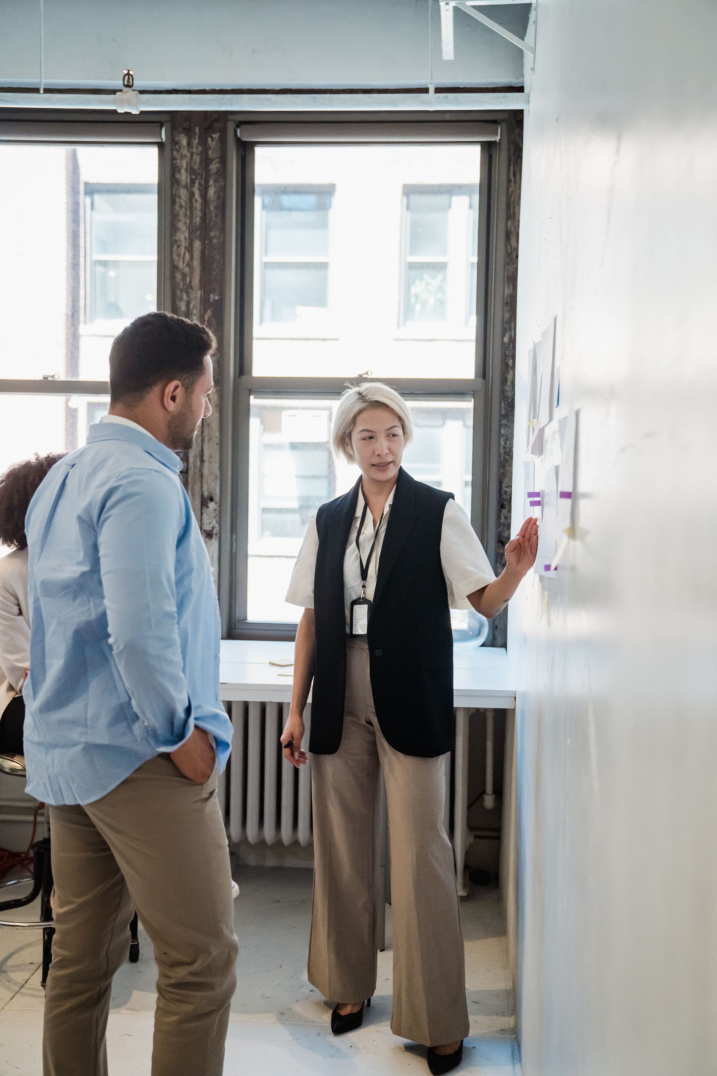 Two men and one woman standing in front of a white wall, engaged in conversation, with a large window in the background.