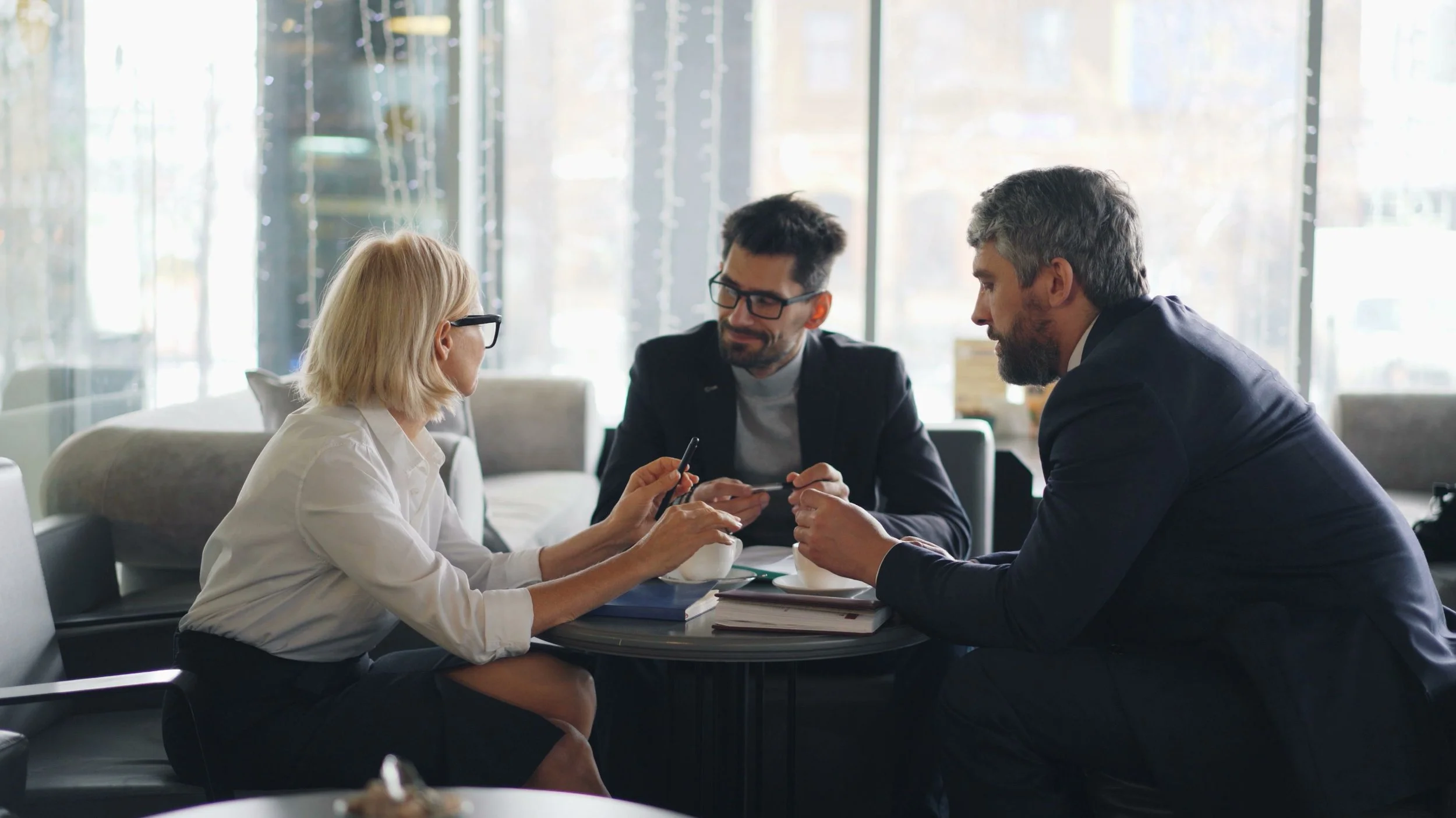 Three people sitting at a table in a brightly lit cafe, engaged in a conversation. Two men are facing a woman, who is showing them something on her phone.