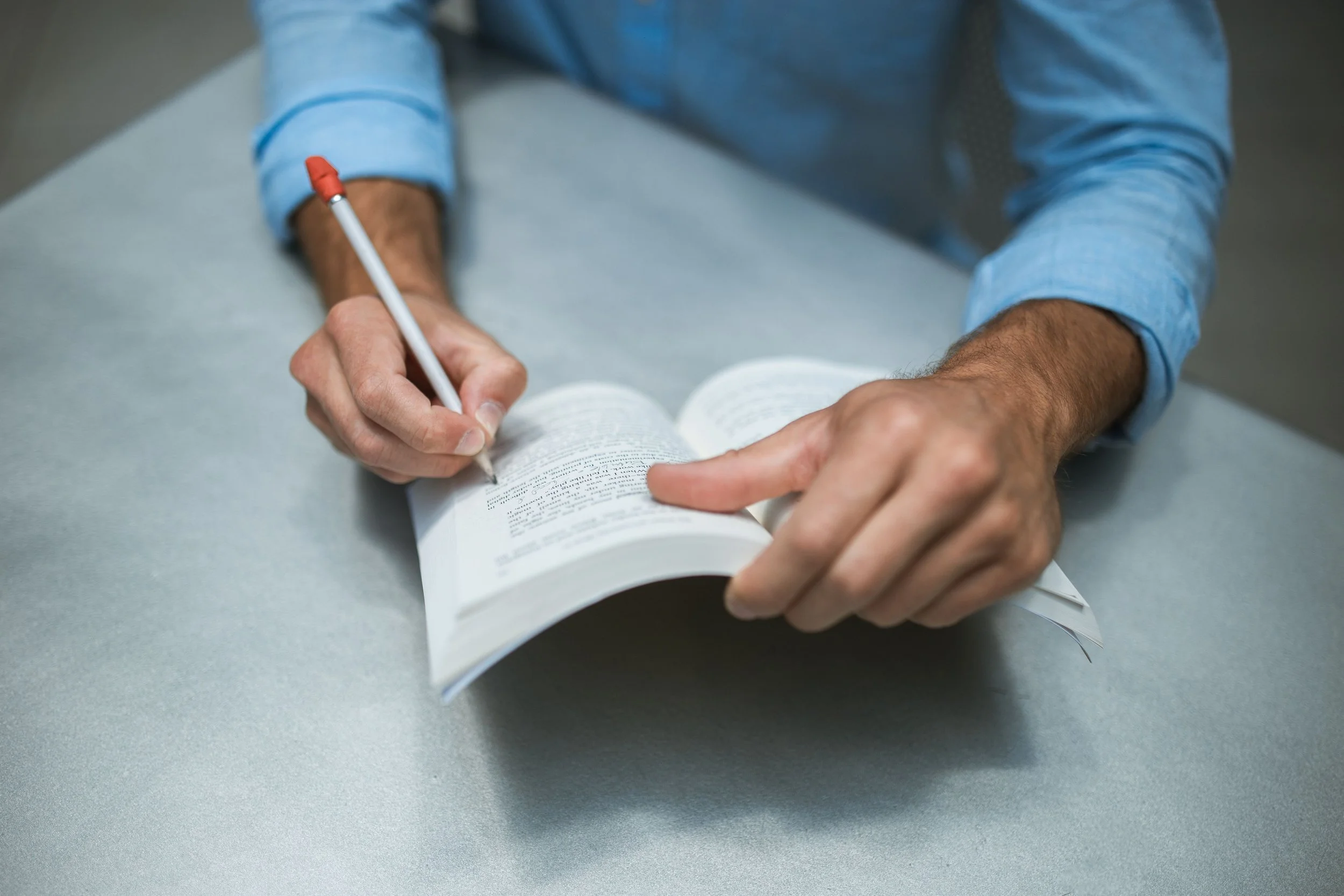 A man wearing a light blue shirt is sitting at a grey table, writing in a book with a red pen.
