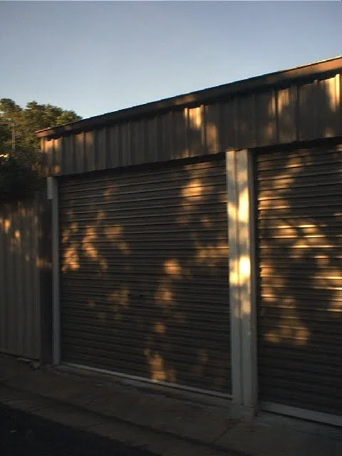 A large metal garage with two closed rolling shutters and a corrugated metal roof, partially illuminated by sunlight with shadows of trees on the shutters.