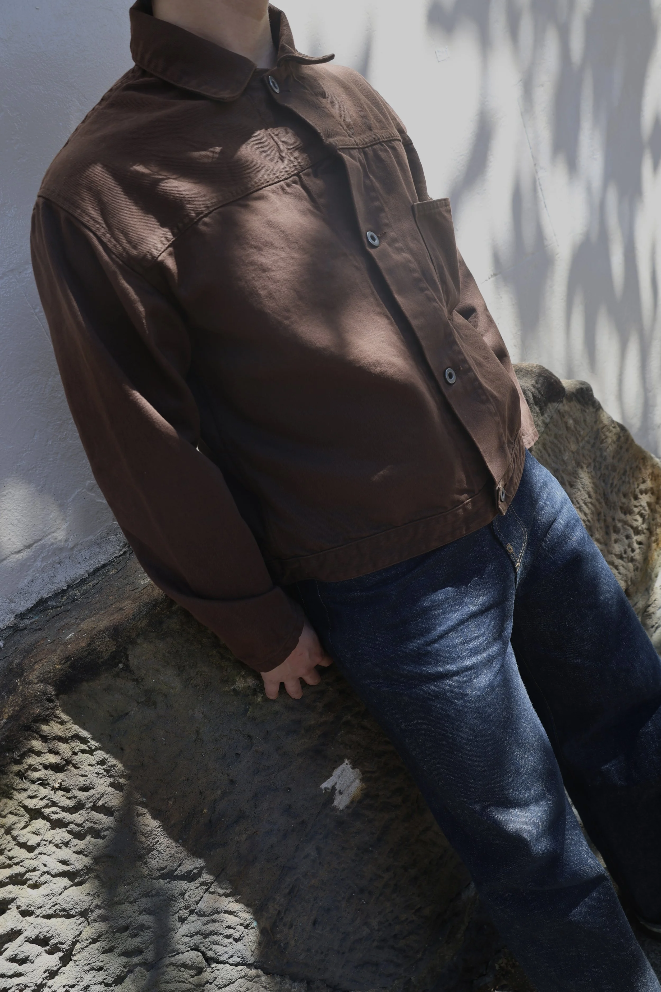 A person wearing a brown jacket and blue jeans leaning against a textured wall, casting shadows on a white wall behind them.
