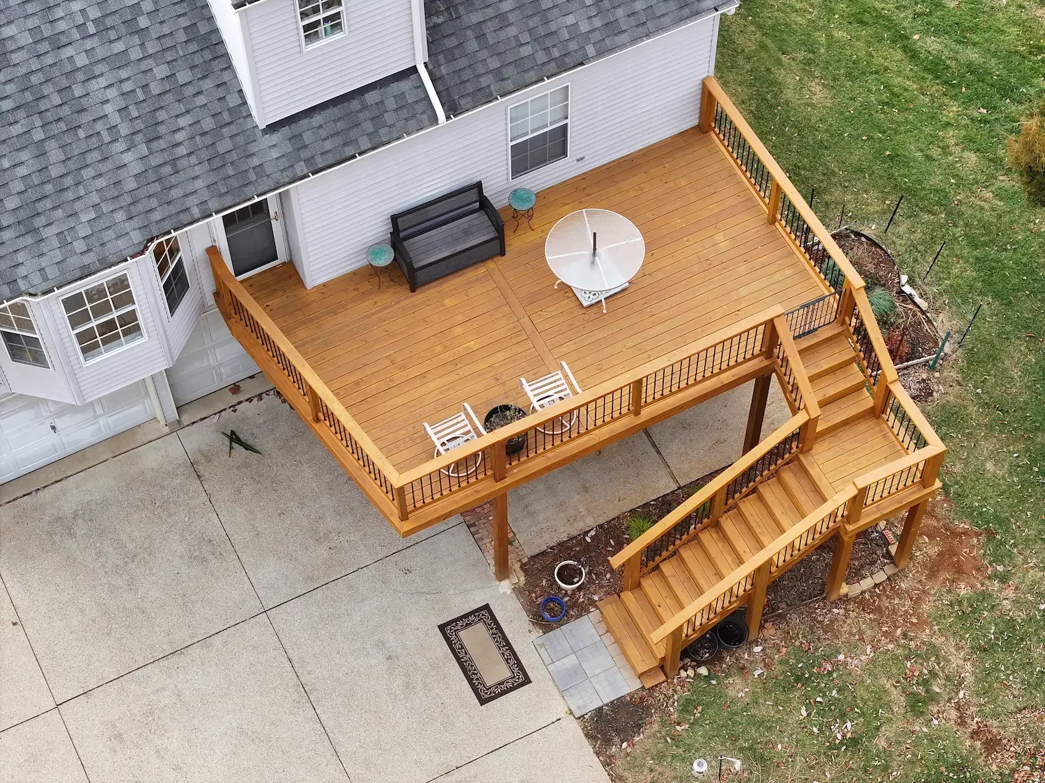 An aerial view of a backyard wooden deck attached to a house, with stairs leading down to a paved area and grass surrounding it. The deck has outdoor furniture, a satellite dish, and some small potted plants.