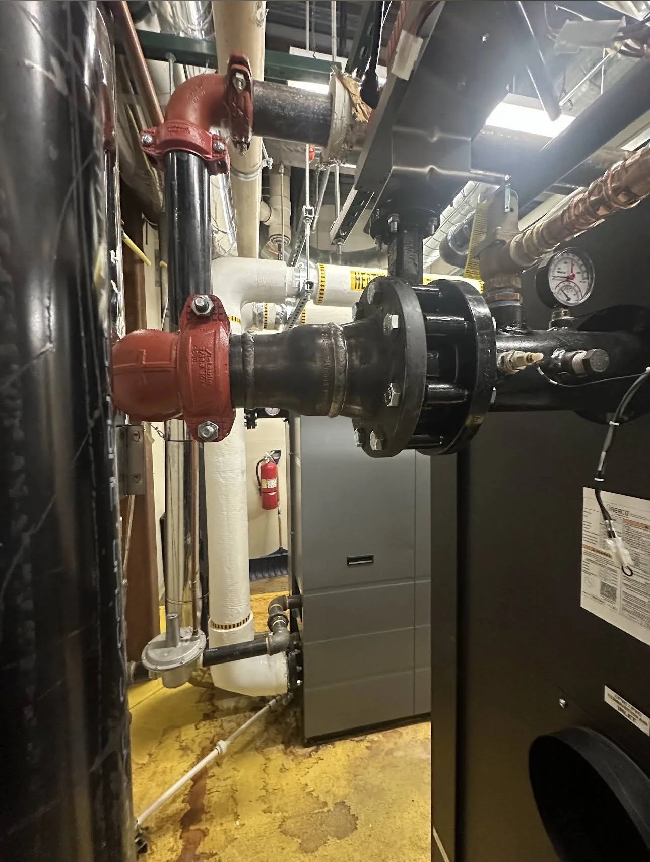 A large pipe with red valve fittings connected to black and white pipes, gauges, and equipment in an industrial or mechanical room.