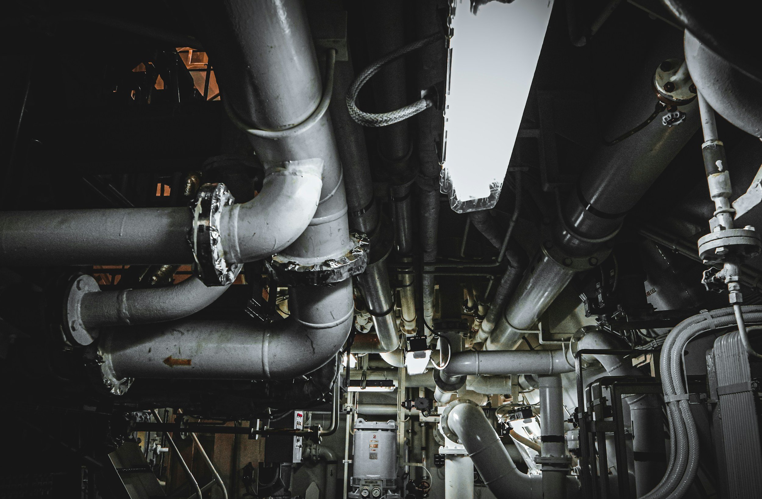 A complex network of industrial pipes and machinery, predominantly gray and metallic, viewed from below.
