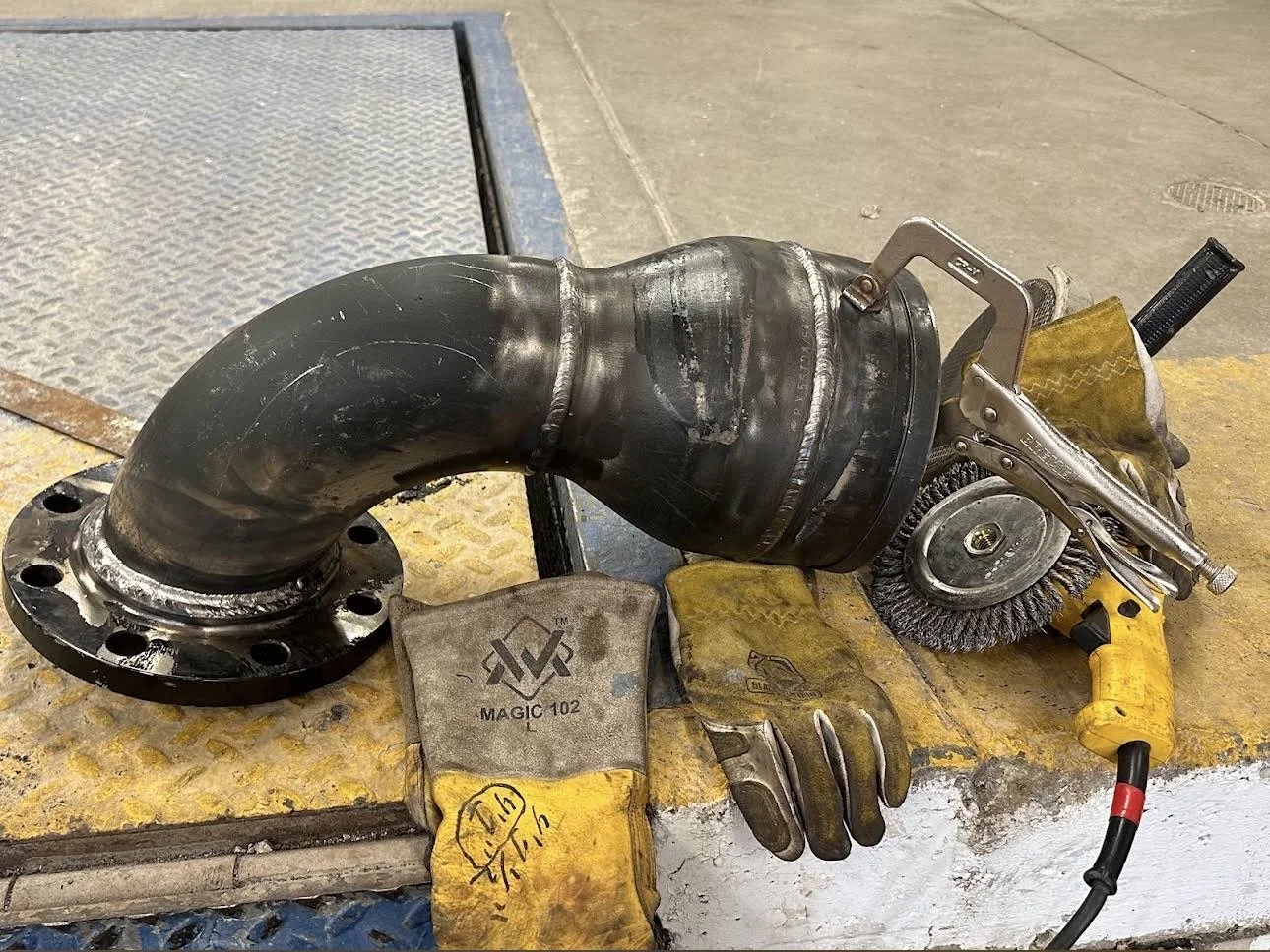 A large black metal pipe with a flange, accompanied by yellow work gloves, a wire wheel brush, and a clamp, on a work surface in an industrial setting.