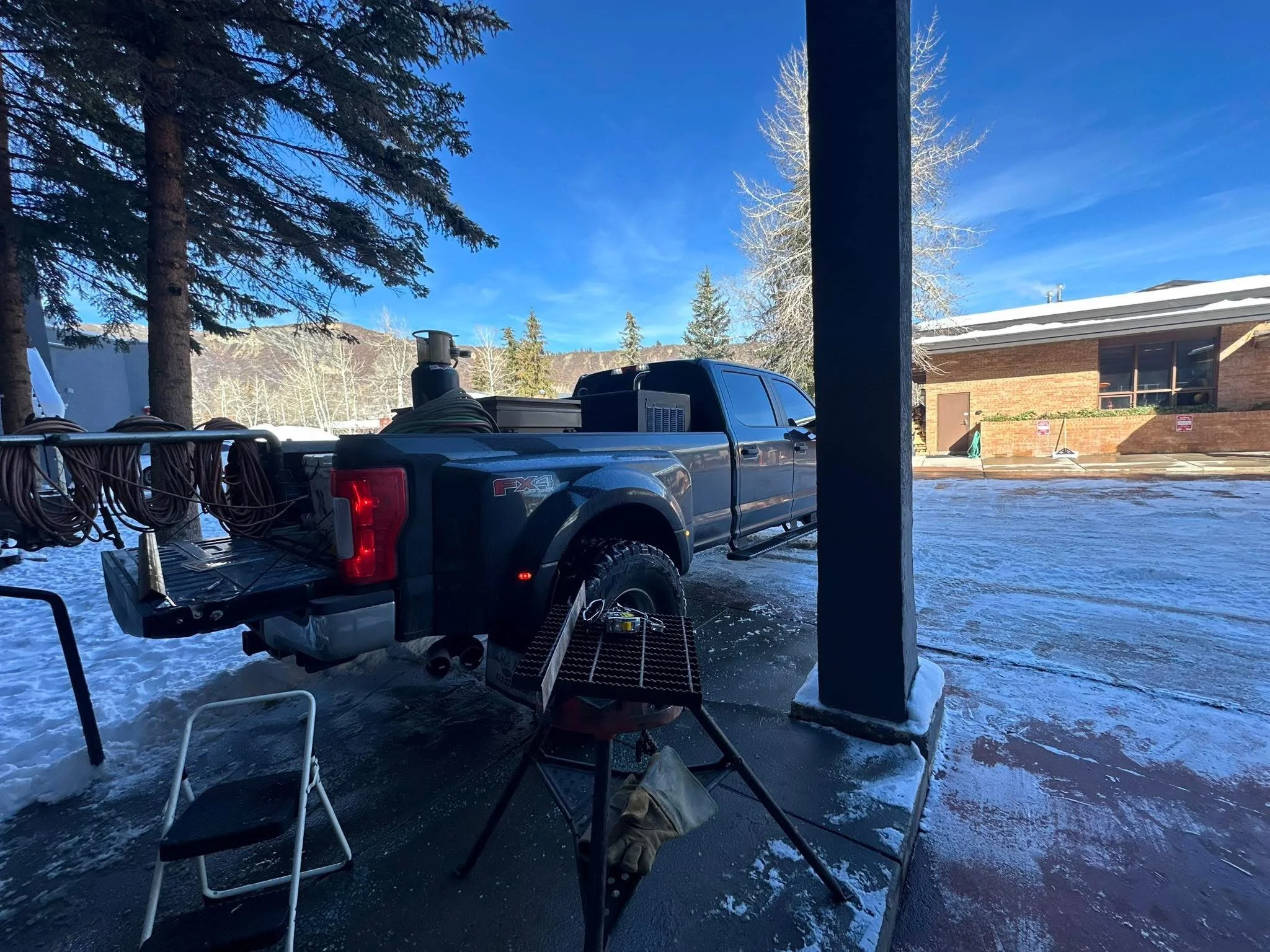 A black pickup truck parked outside a building on a snowy day, with a trailer loaded with hoses or cables attached to it. There are tall trees and a clear blue sky in the background.