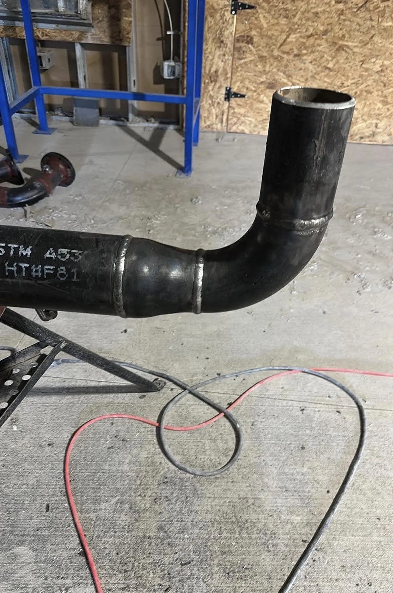 A black industrial pipe with a large open end, connected by welding, placed on a concrete floor in a construction or workshop setting. In the background, there are blue metal framework, electrical boxes, and wooden wall paneling.