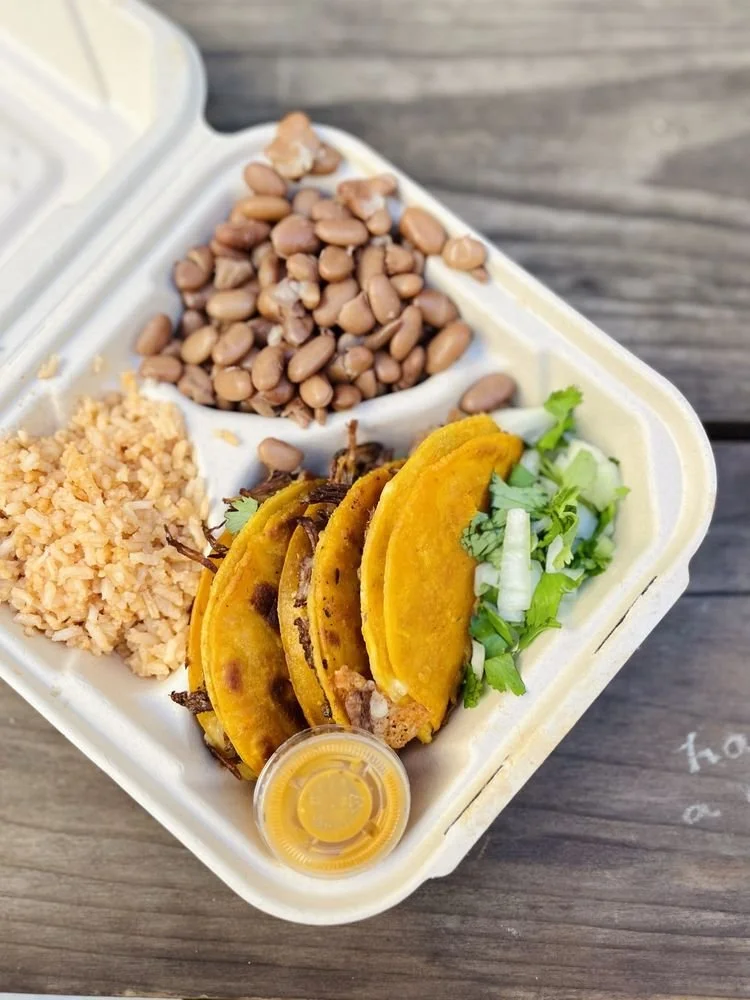 Three tacos with shredded meat, garnished with cilantro and onion, served with rice, beans, and a small container of yellow sauce in a takeout box.