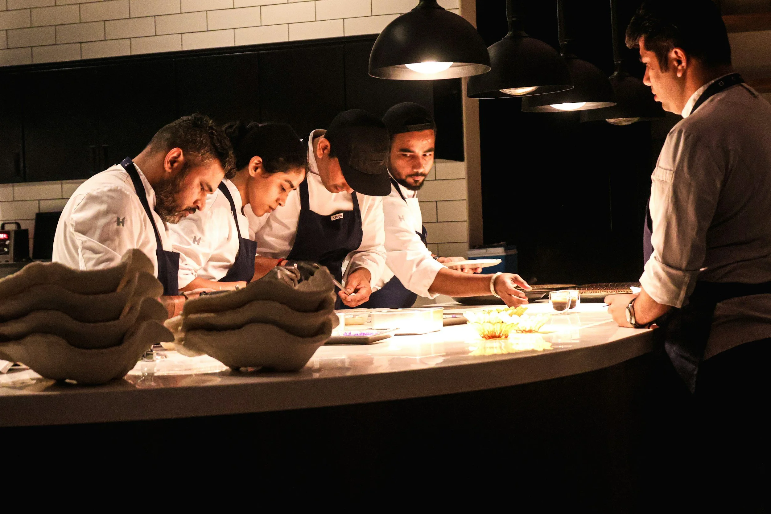 A group of five chefs in white uniforms and black aprons work behind a counter in a dimly lit kitchen, with a single chef on the right facing them, preparing food under hanging lights.