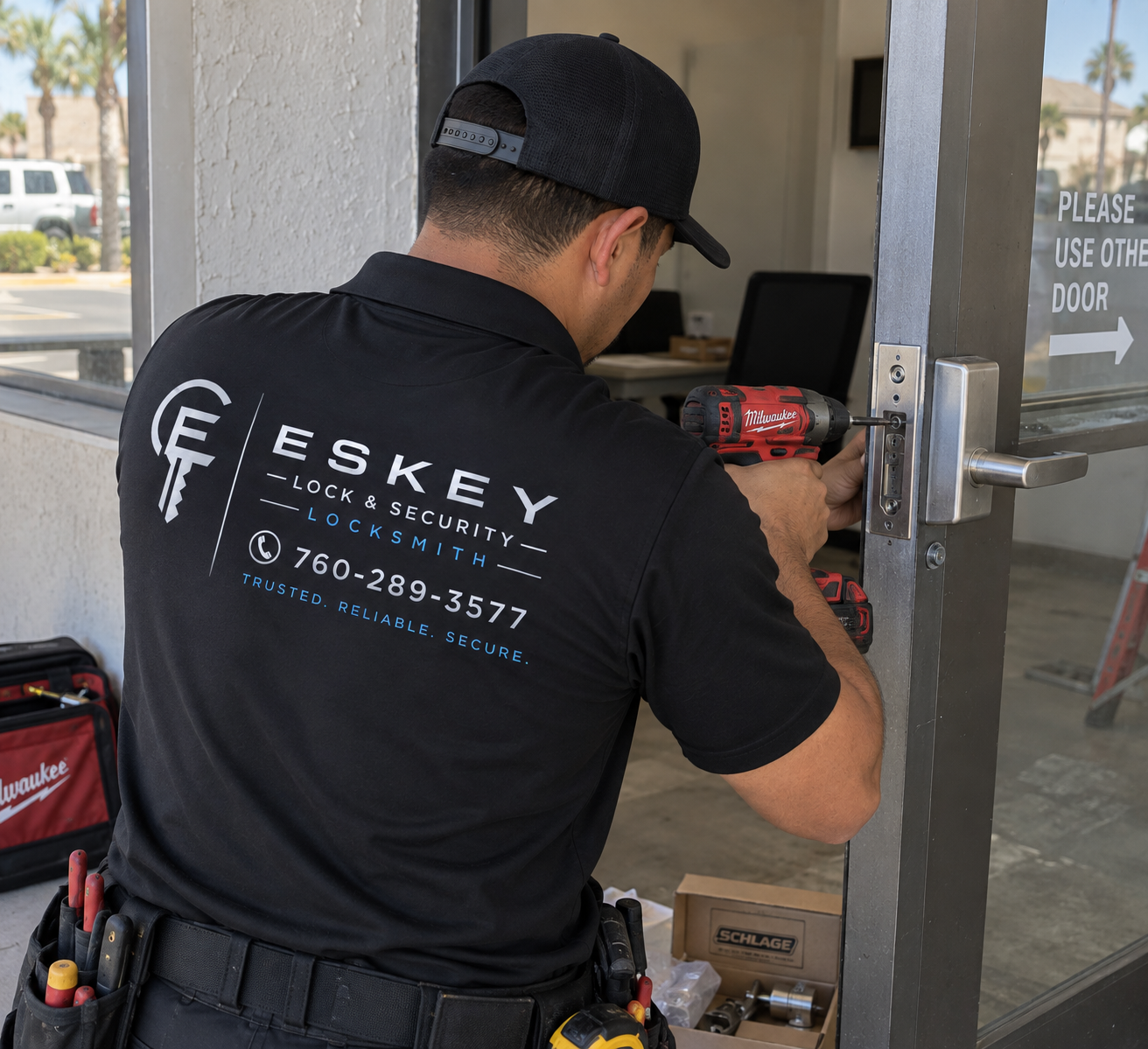 A locksmith working on a door lock with a red power drill, wearing a black shirt with a company logo and phone number, tools in a belt, and a black cap, outside a building with palm trees in the background.