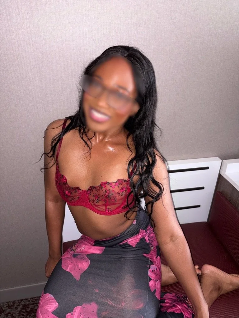 A woman with long black wavy hair kneeling on a red cushioned bench, wearing a red lace bra and a pink and black floral skirt, smiling with her face blurred.