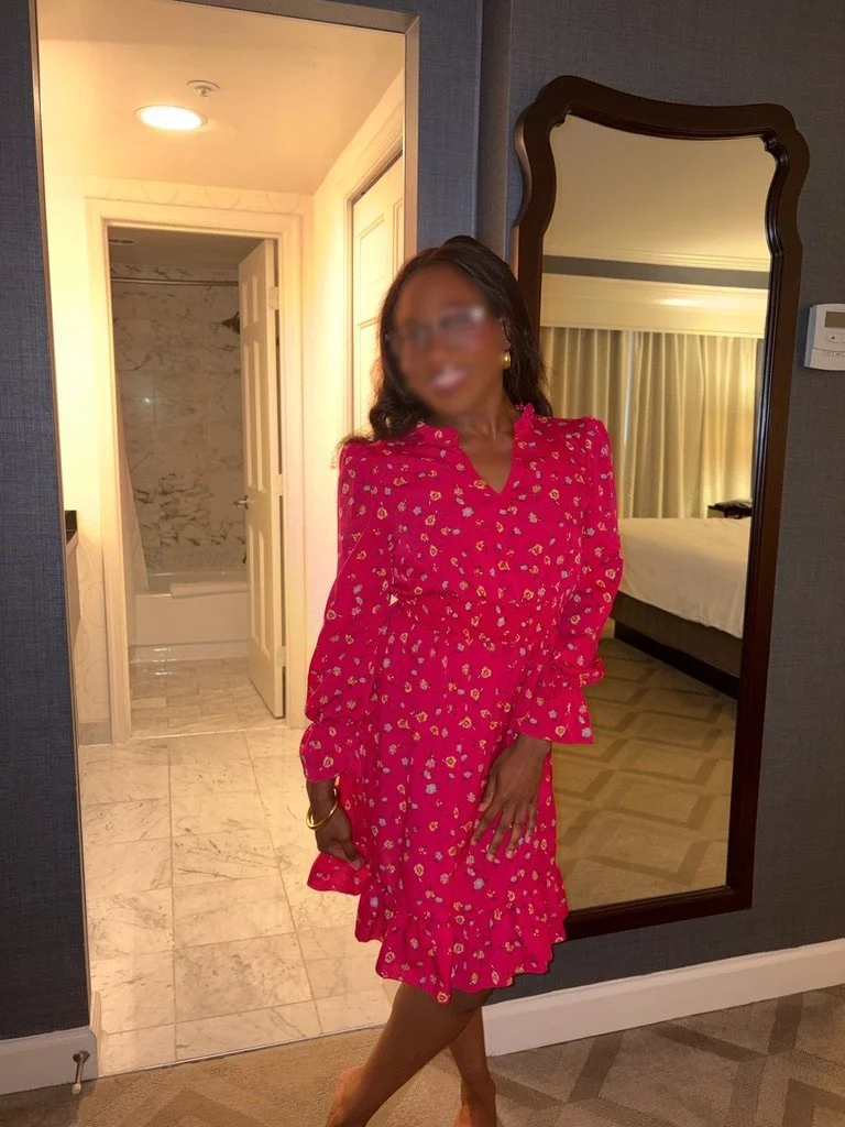 A woman with dark skin and wavy hair wearing a pink floral dress, standing in front of a mirror in a hotel room.