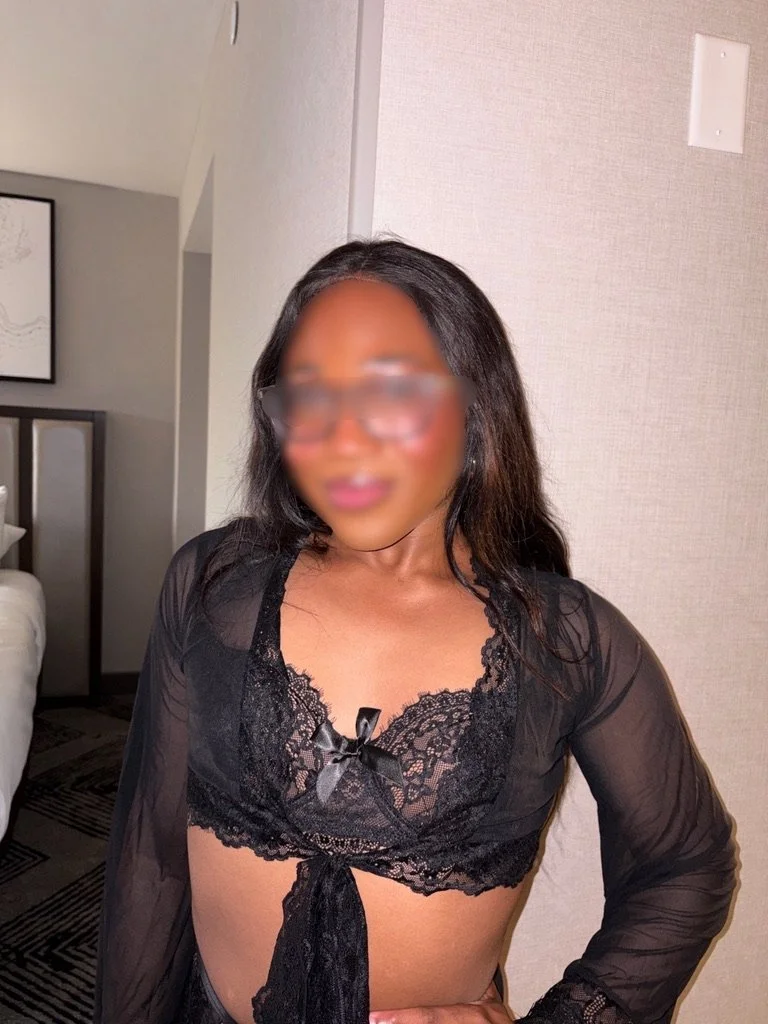 Woman in black lace top with sheer sleeves, standing indoors against a beige wall.