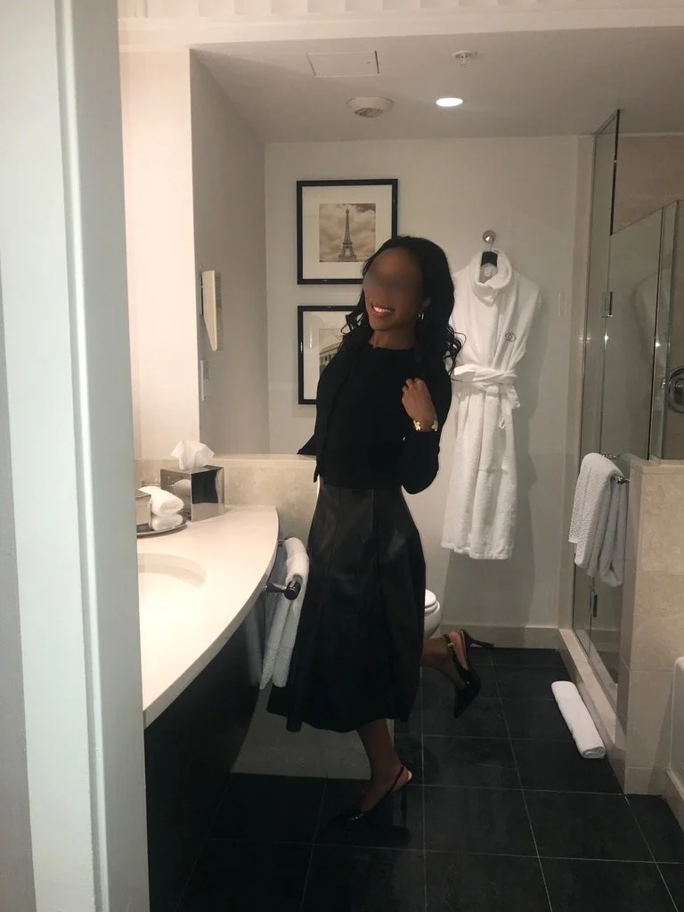 A woman in black outfit with heels standing in a bathroom, smiling at the mirror. There are framed pictures of the Eiffel Tower on the wall, a white bathrobe hanging, and a shower seen in the background.