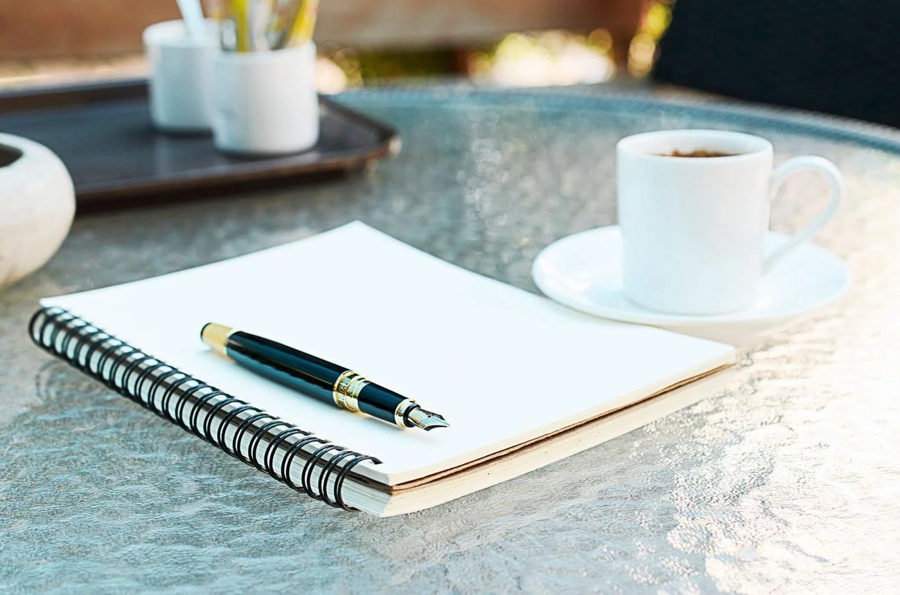 Open notebook with blank page and a fountain pen resting on top, a white coffee cup filled with coffee on a saucer, and a tray with cups and utensils on a textured outdoor table.