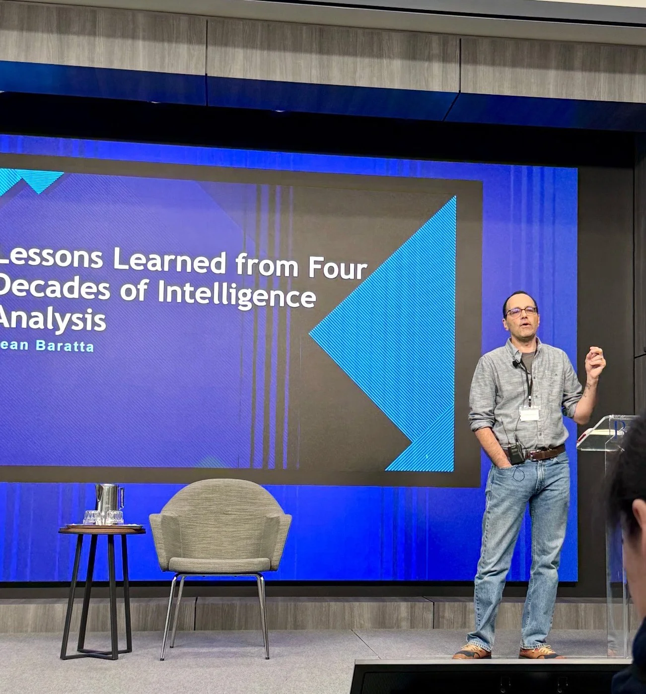 Dean Baratta giving a presentation in front of a large screen displaying the title 'Lessons Learned from Four Decades of Intelligence Analysis'