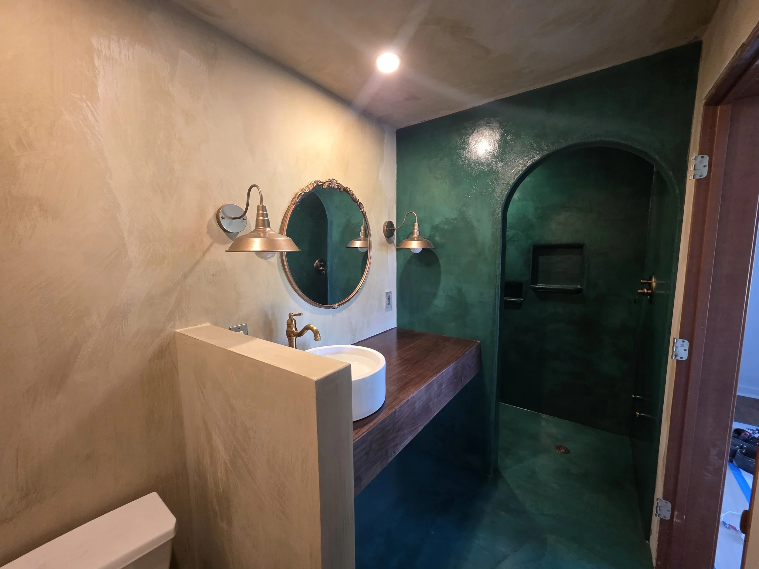Bathroom with a beige textured wall, green accent wall, round mirror, brass wall lights, a white sink, and a wooden vanity counter. An open wooden door is visible on the right.