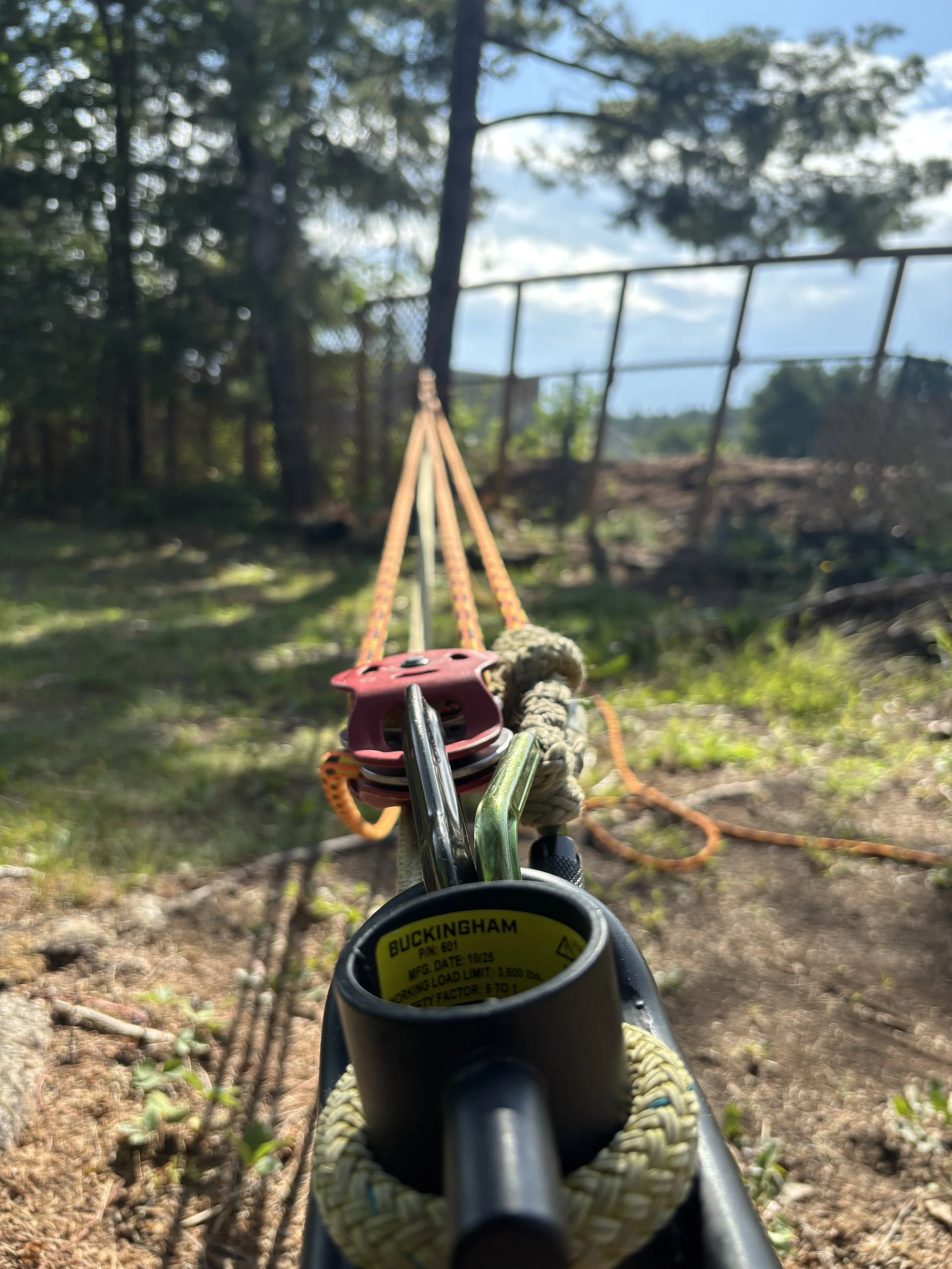 View from the bottom of a tree climber or angle grinder, looking along a rope or cable toward a wooded area with trees, a fence, and cloudy sky in the background.