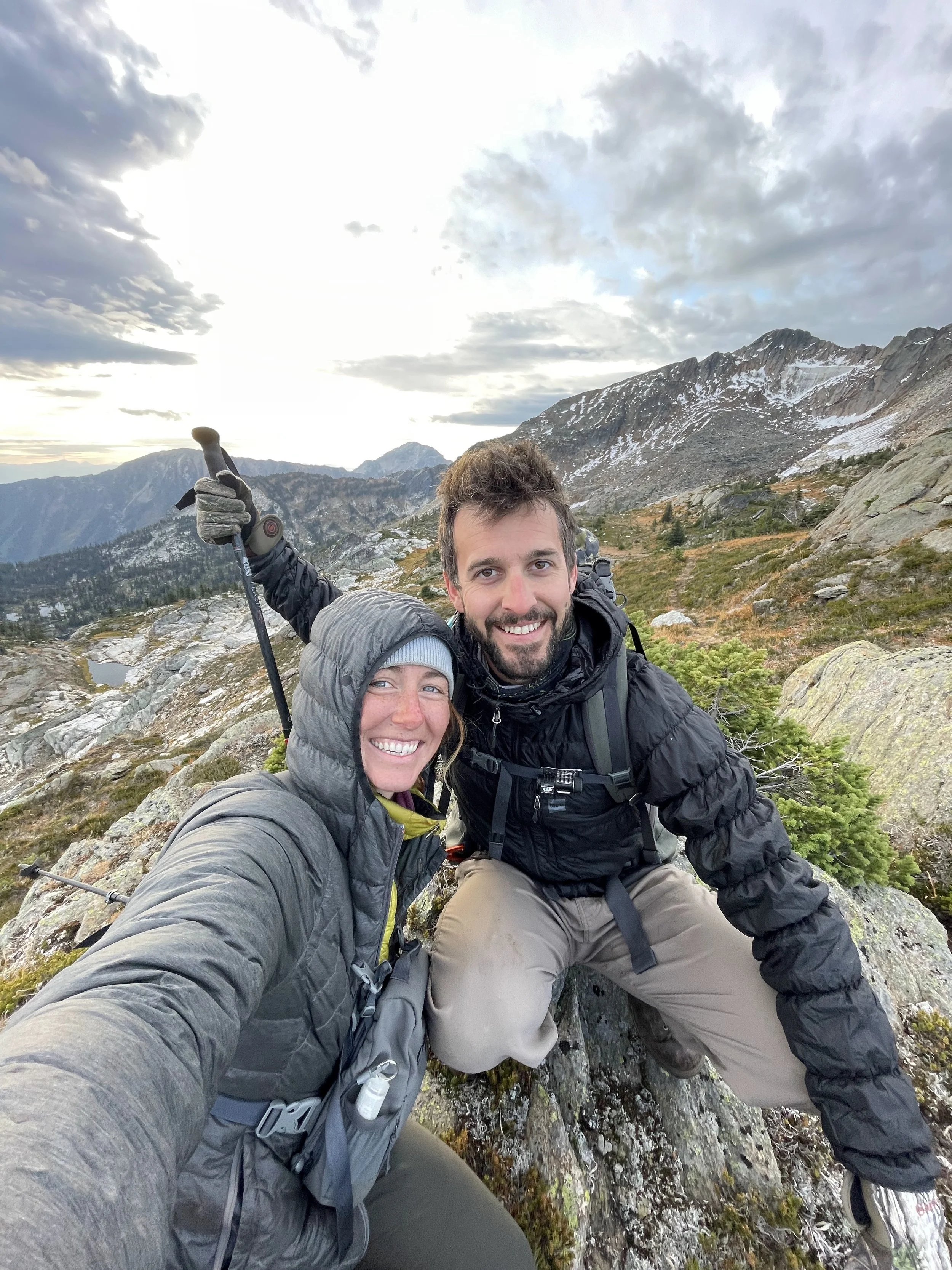 Two hikers, a man and woman, taking a selfie on a mountain trail with a scenic mountain range and cloudy sky in the background.