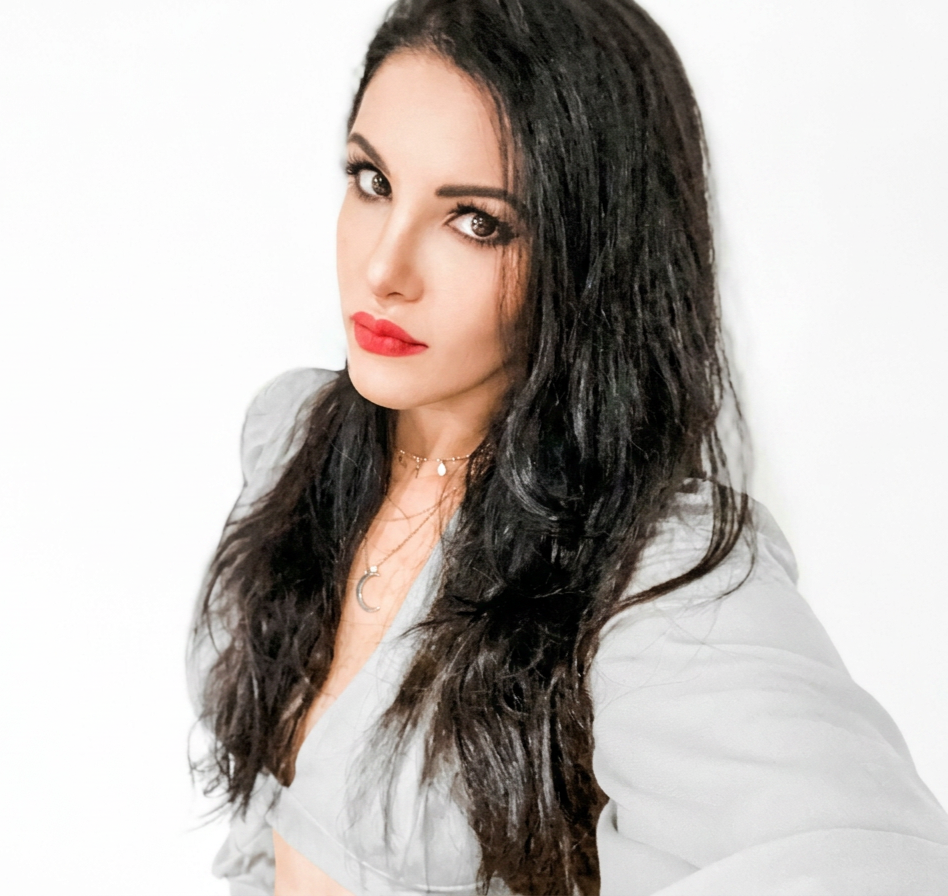 A woman with long, wavy black hair and red lipstick is taking a selfie against a white background. She is wearing a gray blazer and layered necklaces.