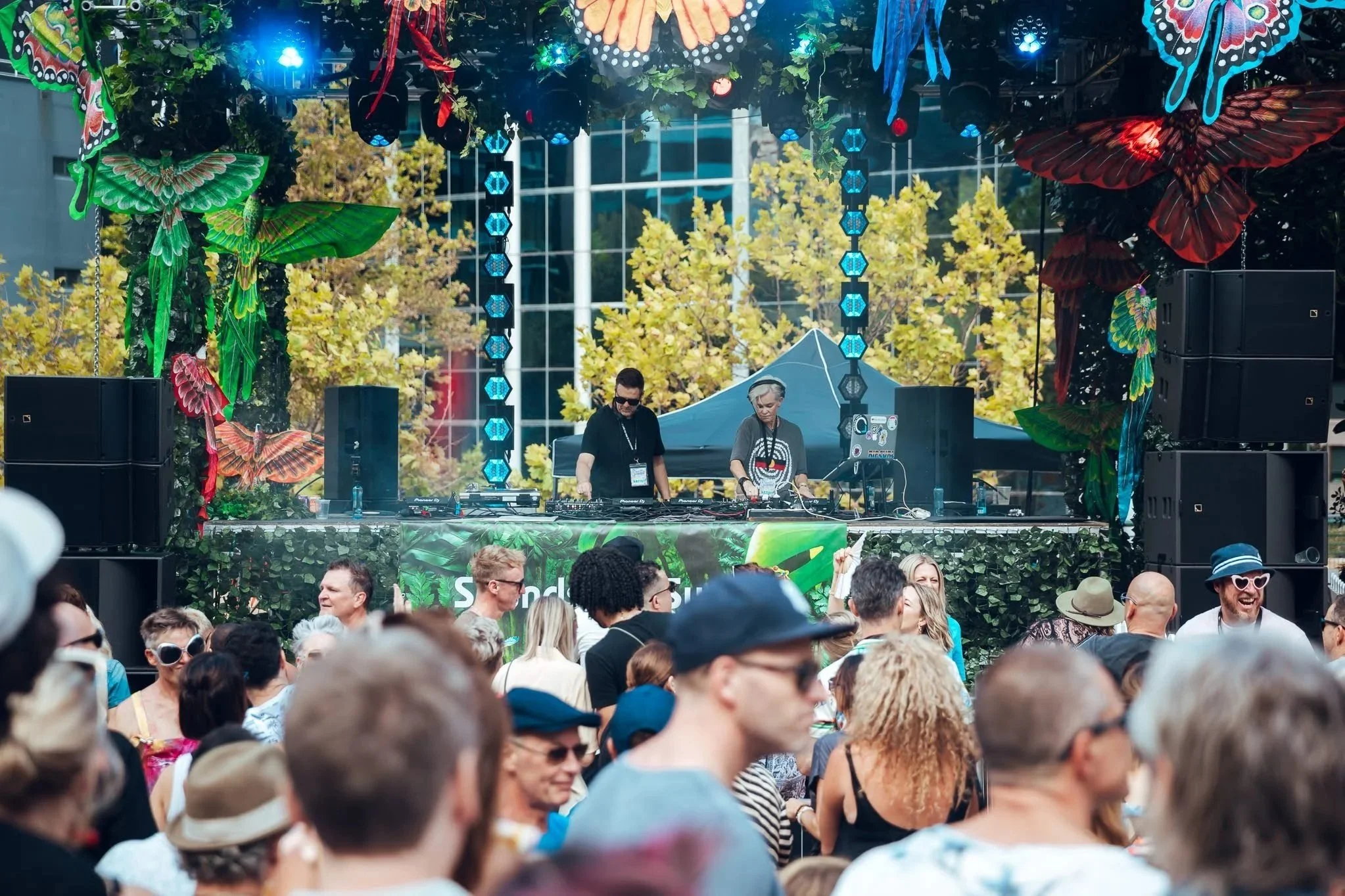 Dance music performance on an outdoor stage with colorful butterfly decorations, a DJ setup, and a crowd of people enjoying the event during daytime.