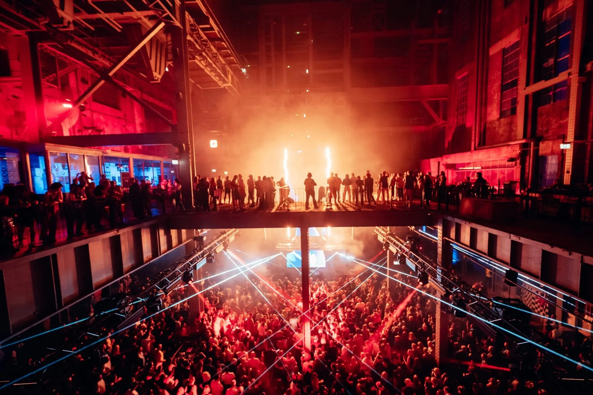 Crowd of people at a concert or nightclub with red lighting and laser lights, inside a large industrial-style building with exposed brick walls and metal structures.