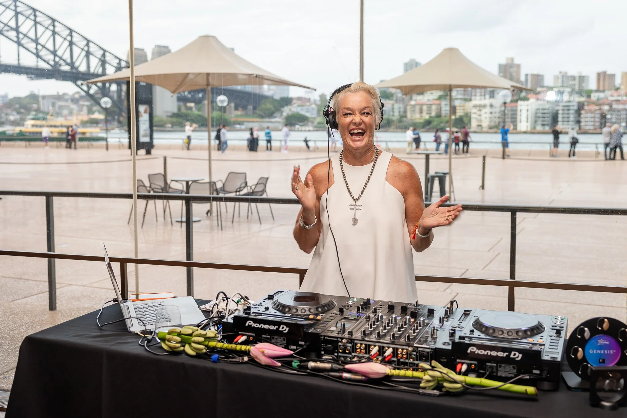 DJ Kate Monroe wearing a white sleeveless top, headphones, and a necklace is smiling at a DJ setup outdoors with a city skyline and water in the background. The DJ equipment includes a Pioneer DJ controller and a laptop on a black table.