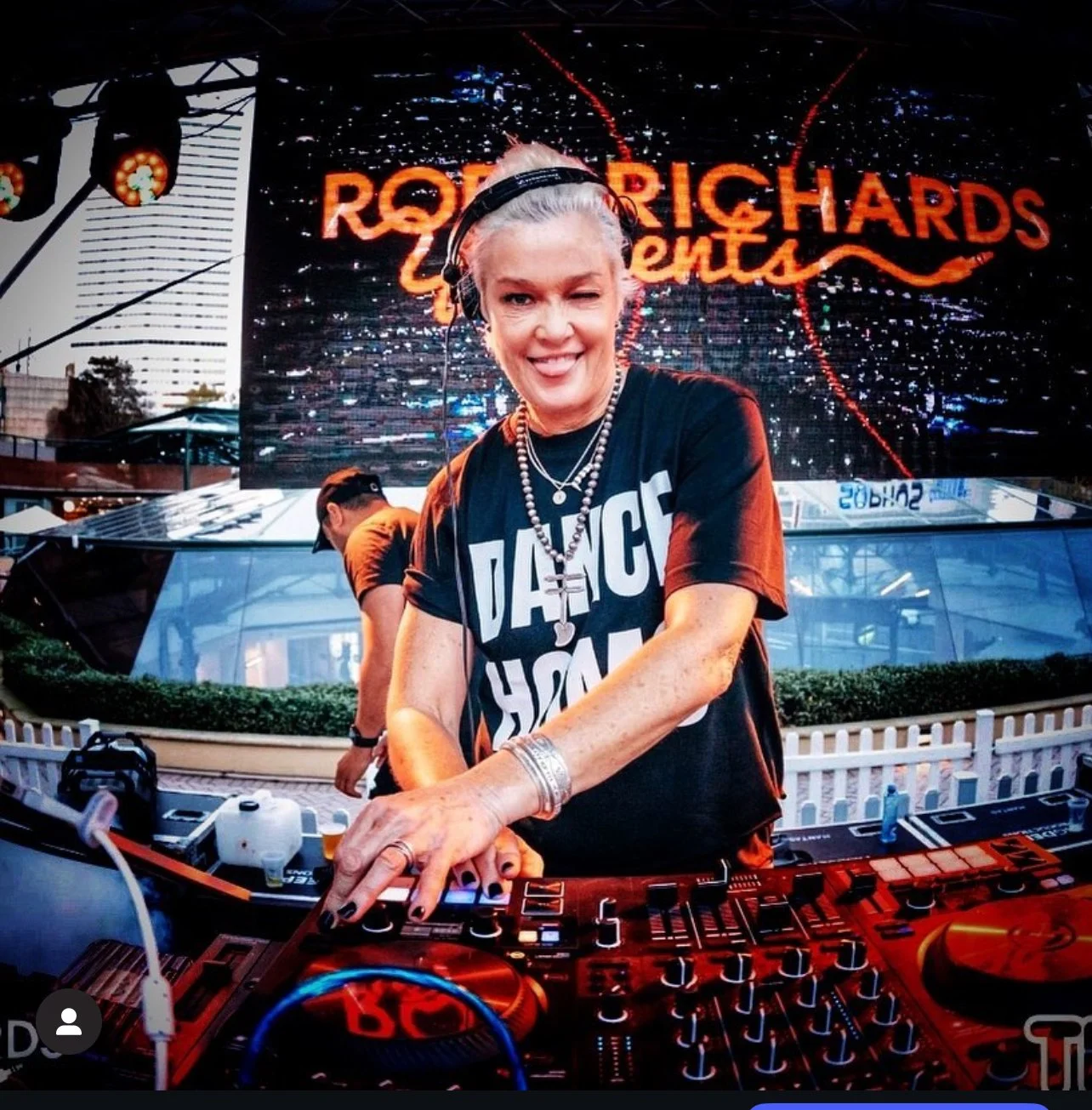 DJ Kate Monroe, wearing a black T-shirt that says dance hall, and jewelry, is smiling and winking while working at her DJ booth with a large LED screen behind her.