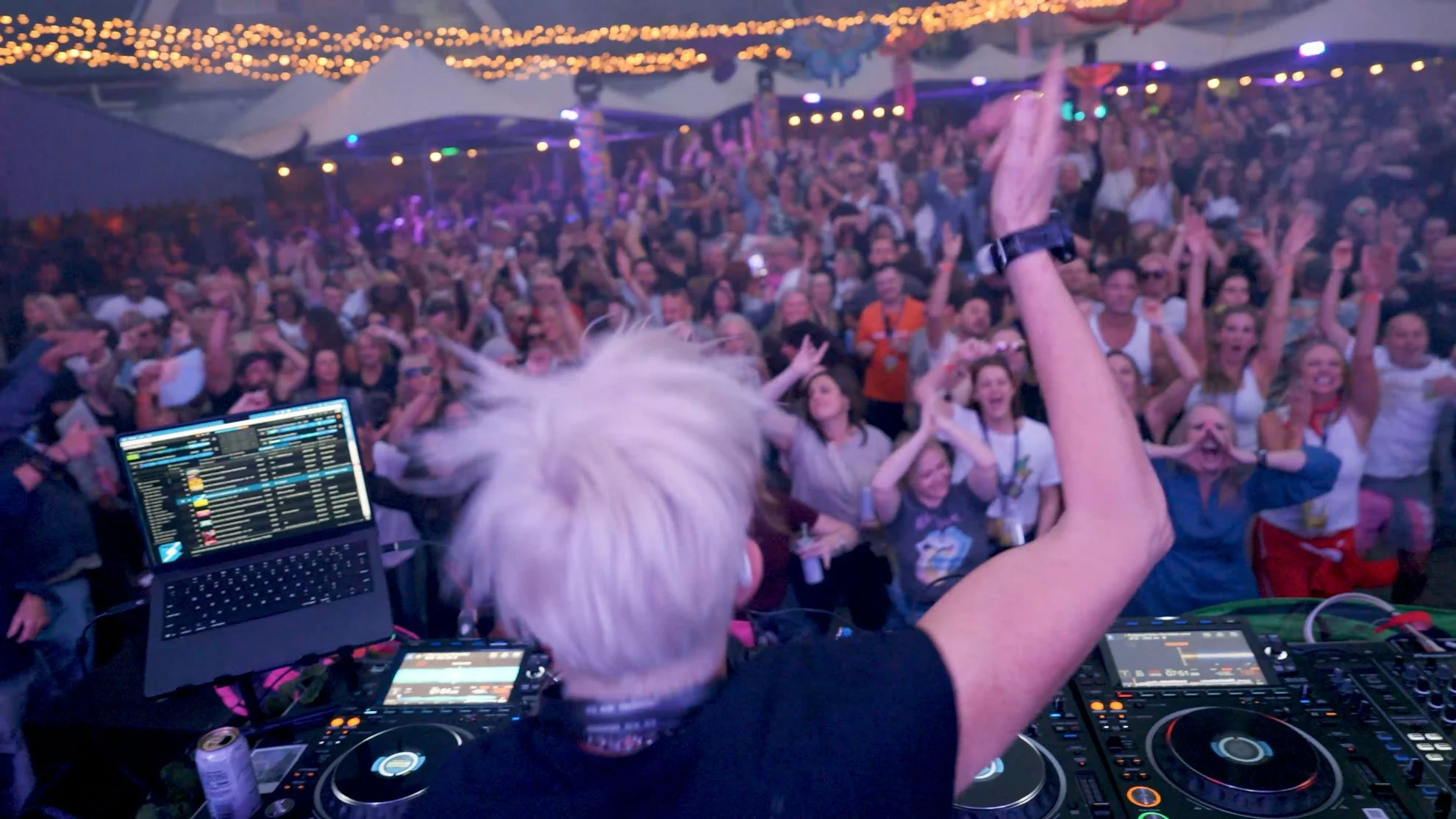Crowd of people dancing and enjoying a music event under string lights, with a DJ performing on stage in the foreground.
