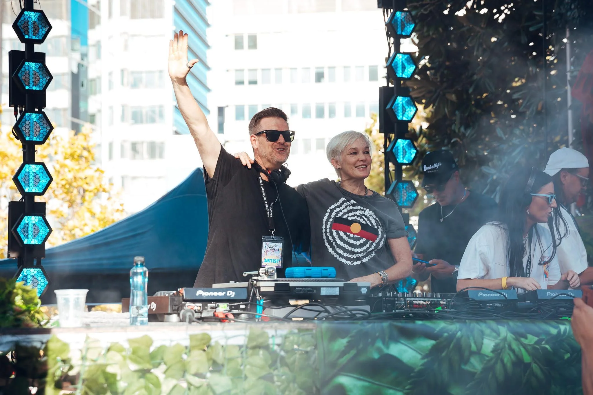 Two people, a man and a woman, are standing behind a DJ booth at an outdoor event. The man wears sunglasses and waves, while the woman smiles. There are other people in the background and large buildings behind them.