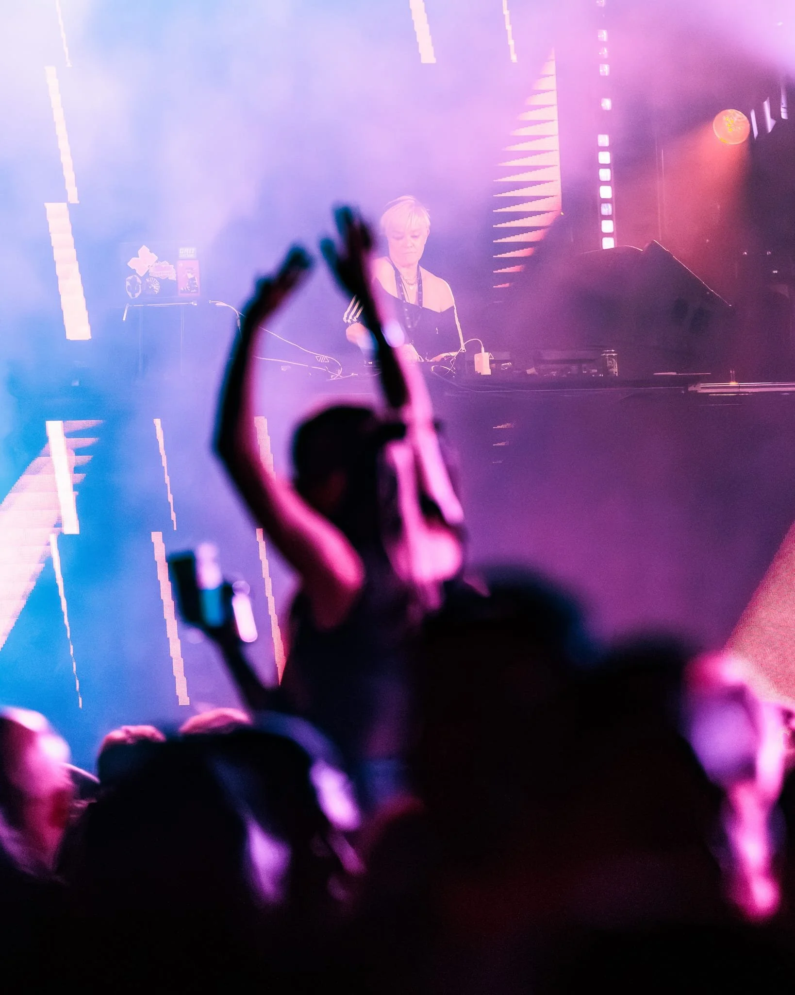 A crowded concert scene with DJ Kate Monroe performing on stage, purple and pink lighting, an audience with hands raised, and fog or smoke effects.