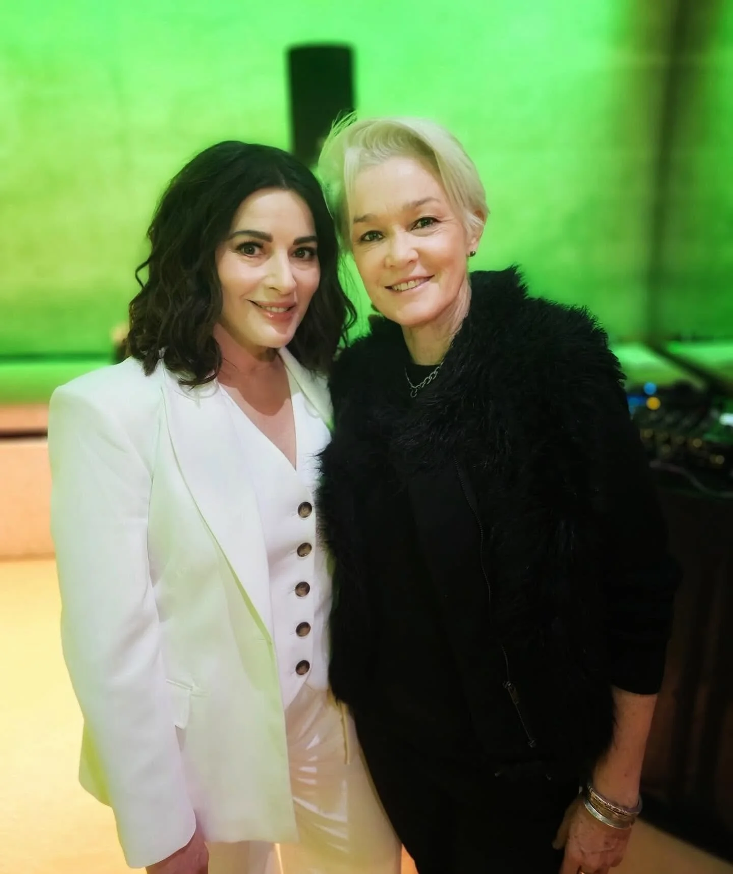 Two women standing close together and smiling, with a green background.