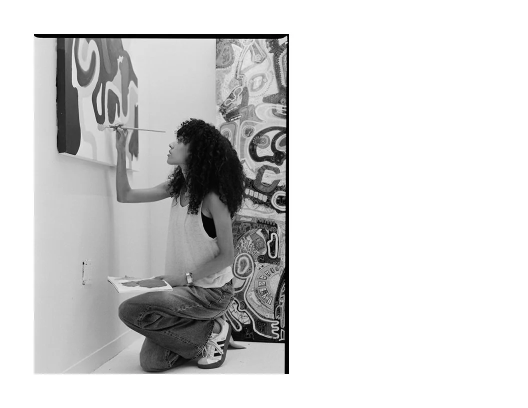 A woman with curly hair kneeling on the floor, painting or drawing on a wall with a paintbrush, holding a palette in her left hand, with abstract artwork behind her.
