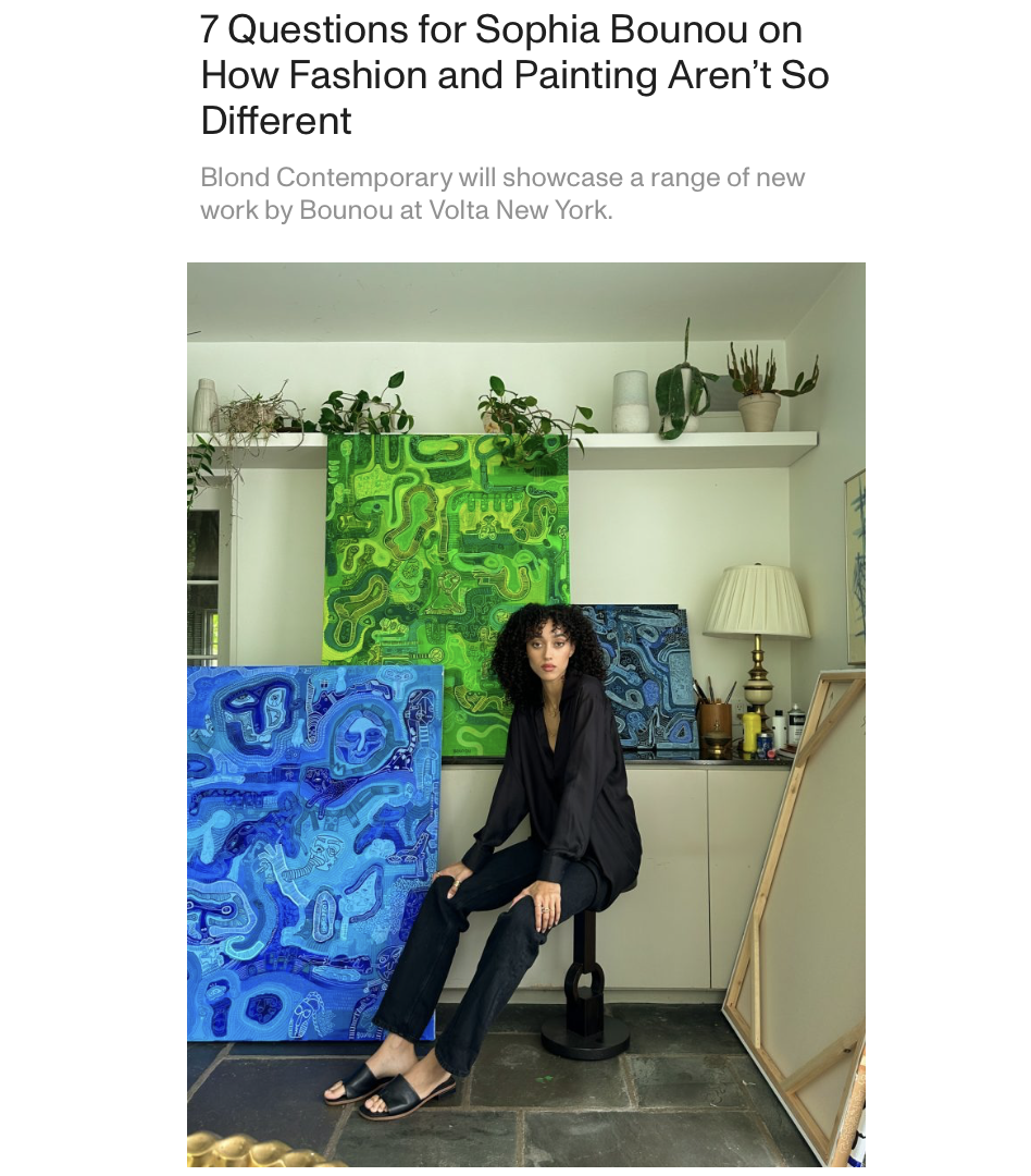 A woman with curly black hair wearing black clothing seated on a stool in an art studio surrounded by colorful abstract paintings and plants.