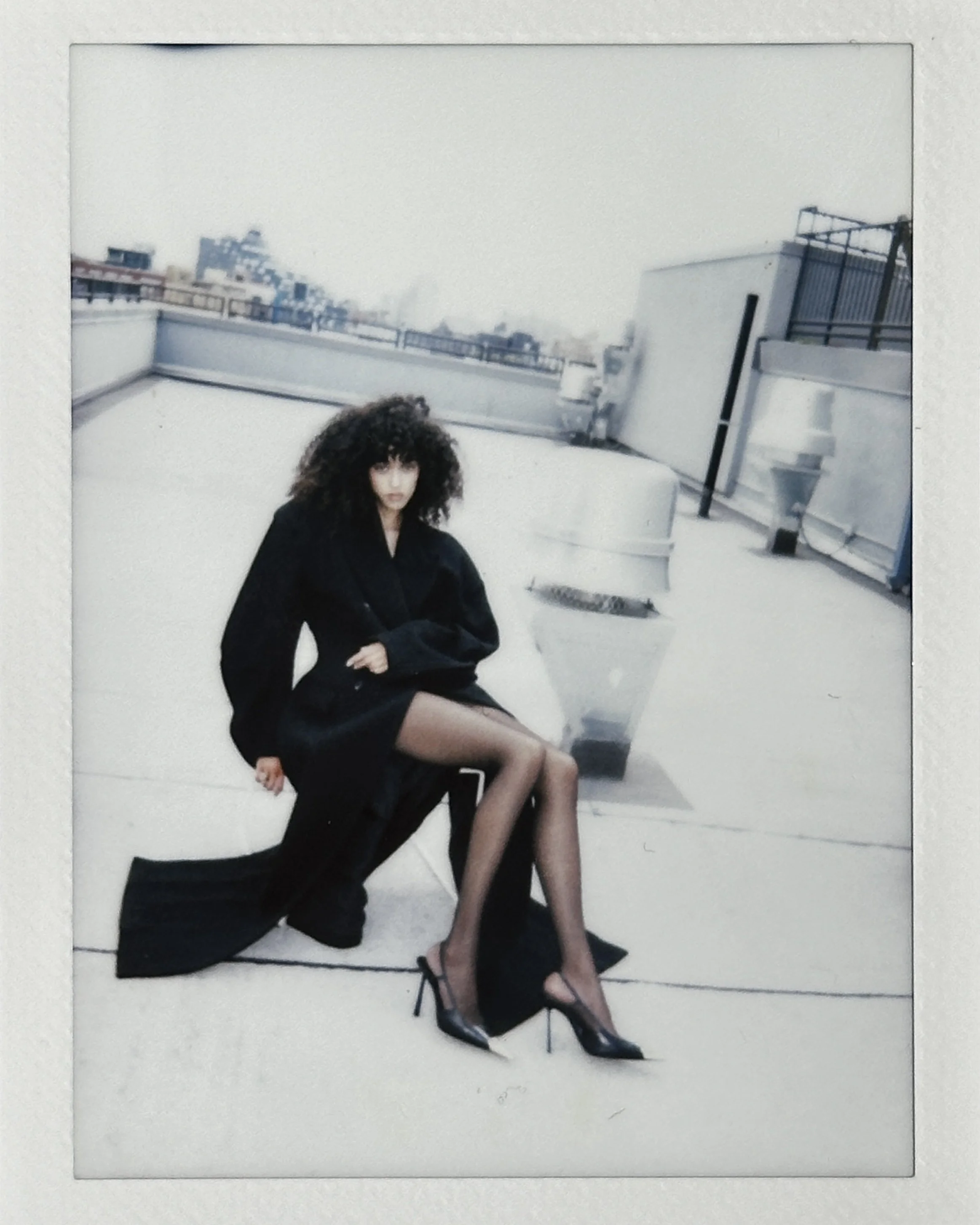 A woman with curly hair wearing a black jacket, black stockings, and high heels sitting on a chair on a rooftop. The background shows a cityscape with buildings and a cloudy sky.