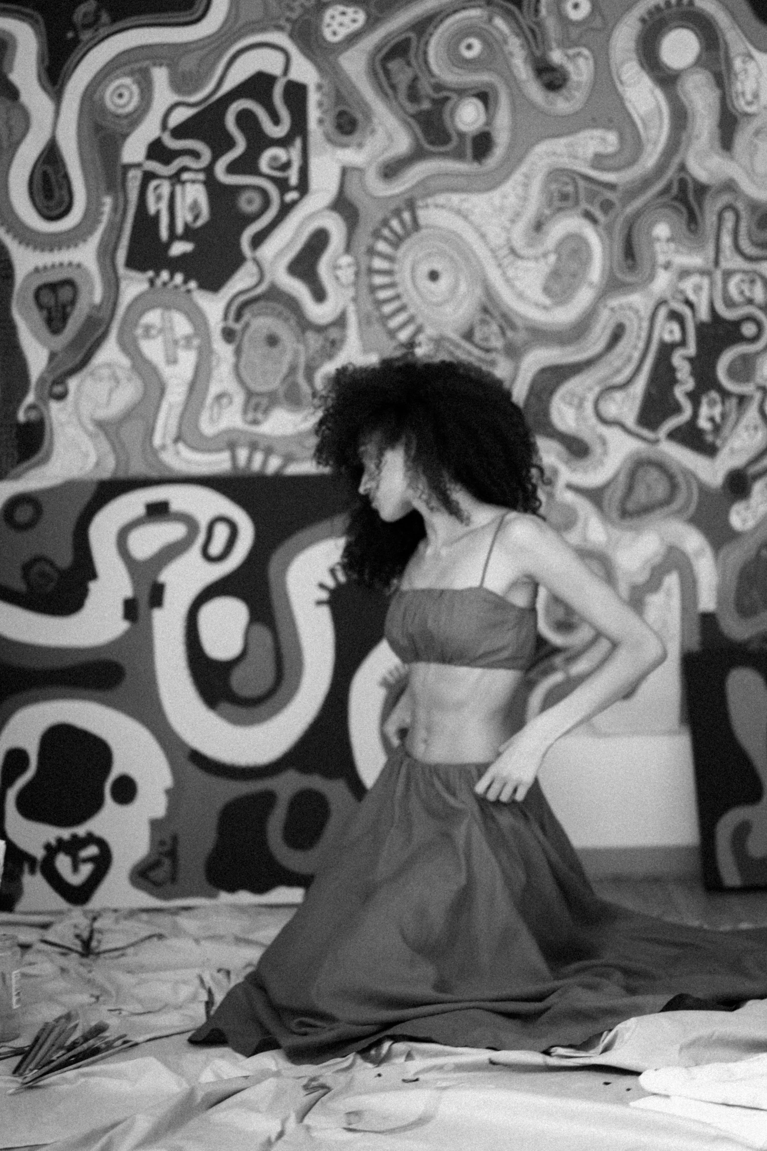 A woman with curly hair wearing a strapless top and skirt sitting cross-legged on a bed with a patterned chintz wallpaper featuring various skulls in the background.