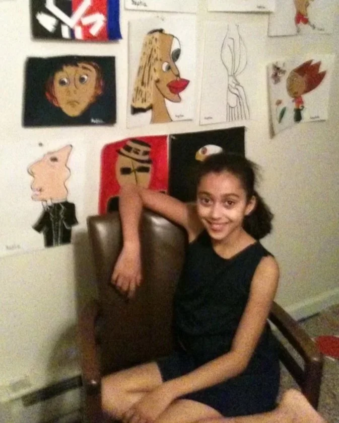 A girl sitting on a chair in front of a wall with various drawings. Her right arm rests over the back of the chair, and she is smiling at the camera.
