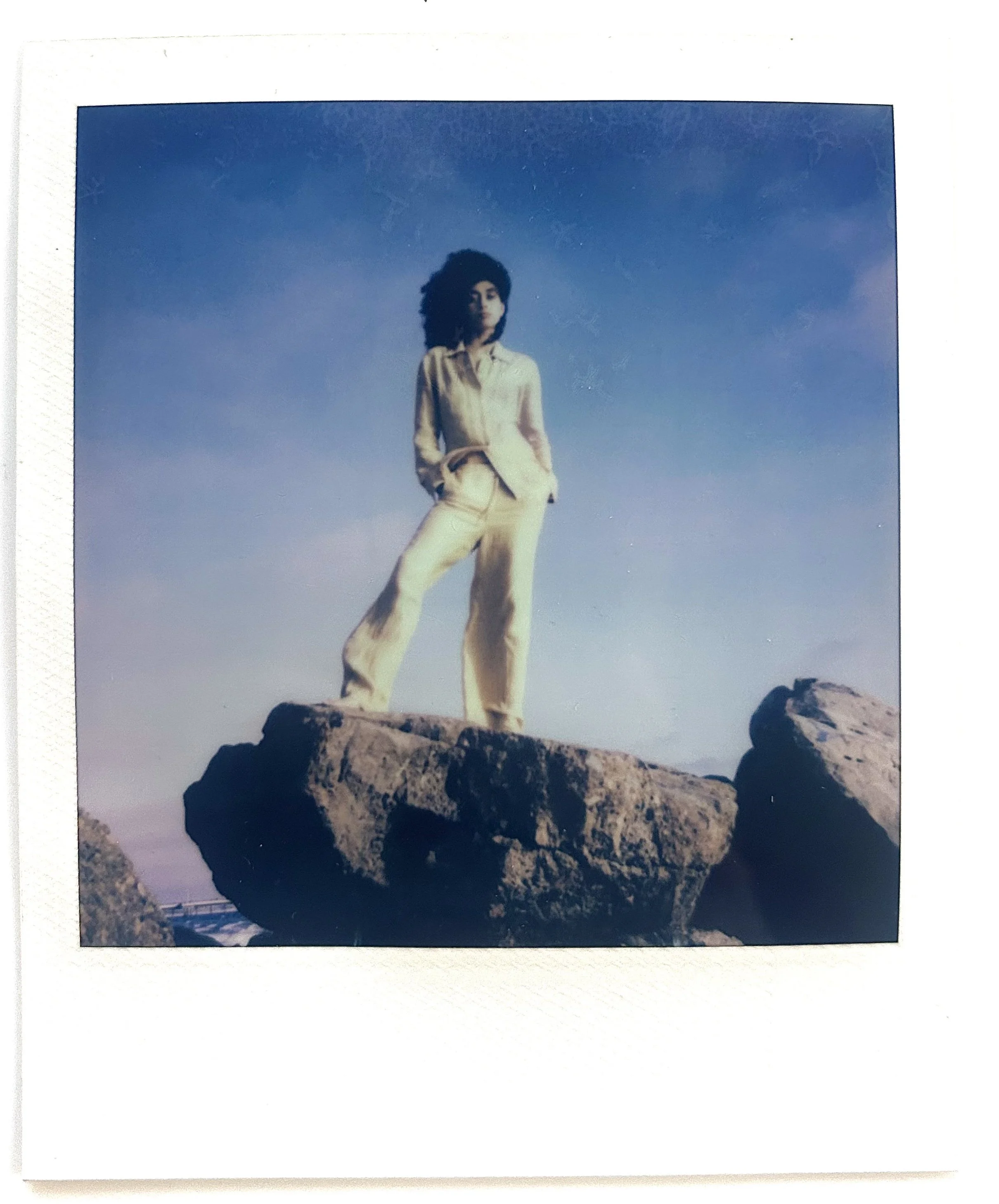 A person with curly hair wearing a light-colored suit stands on a large rock formation against a blue sky.