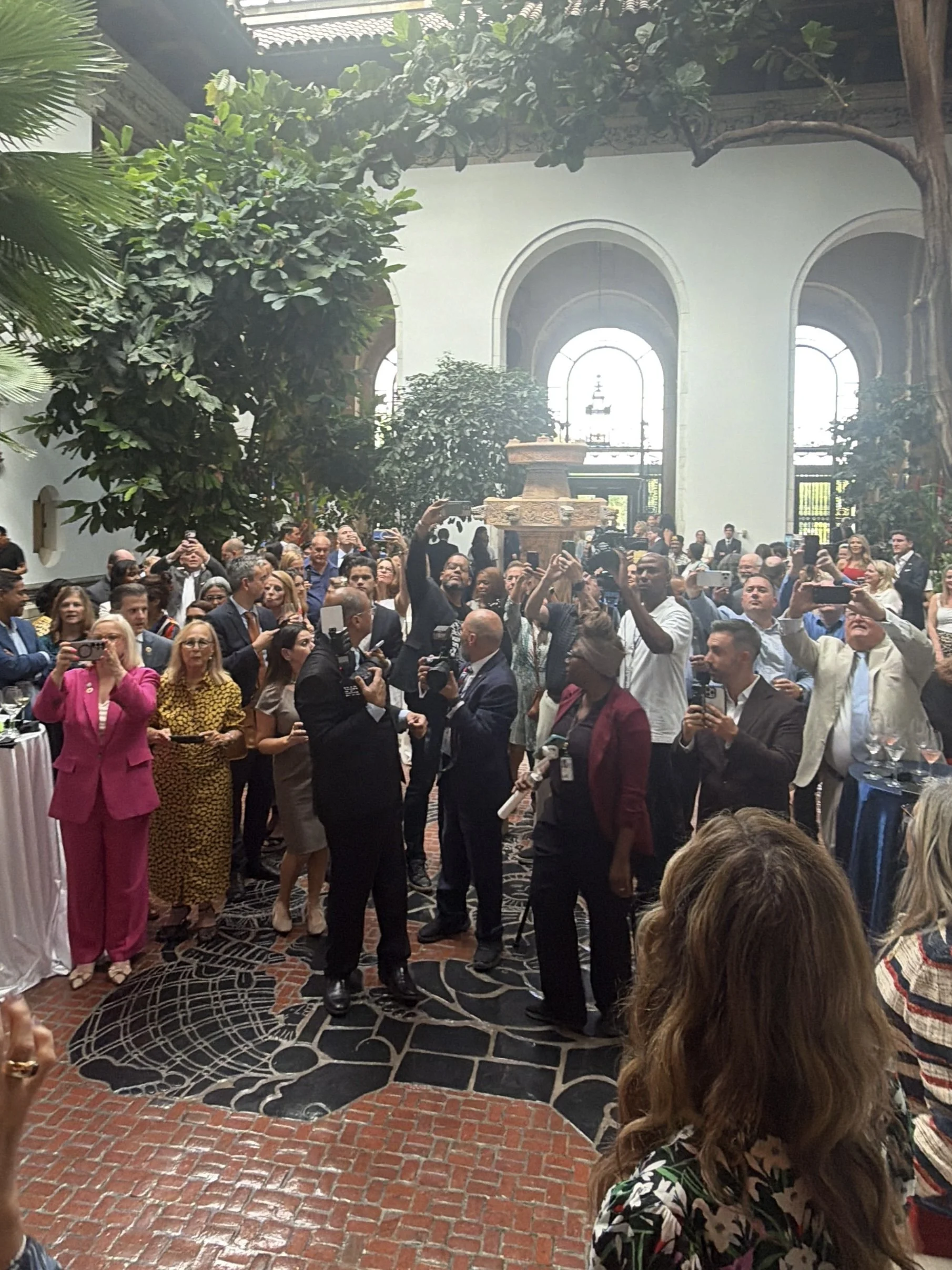 A large group of people in formal attire gathered in a spacious, elegant room with tall arched windows and ornate decorations, taking photos and engaging in conversations.
