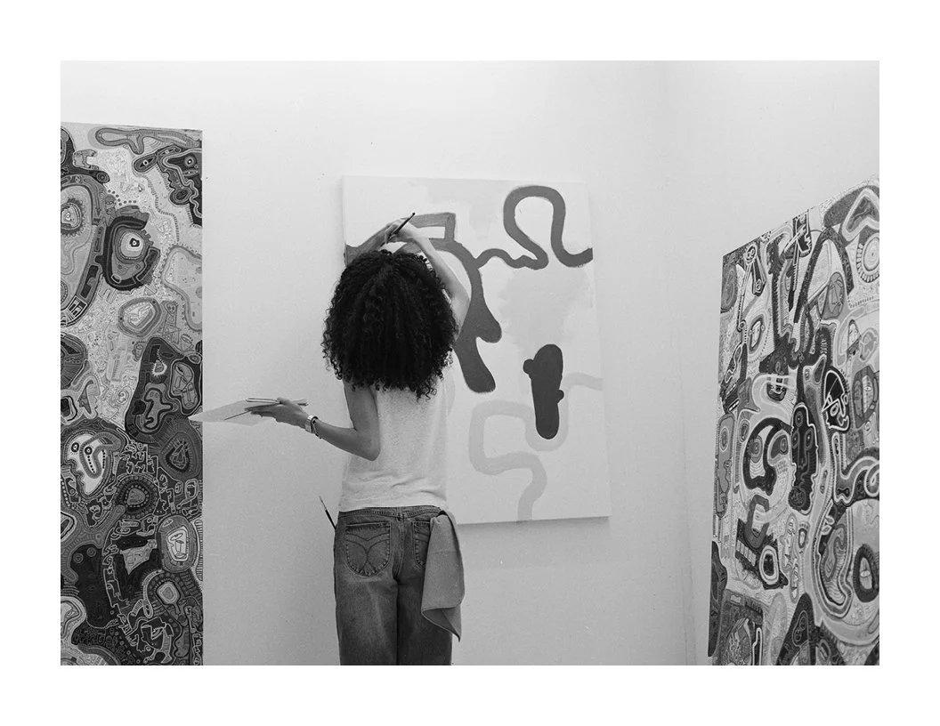 A woman with curly hair painting abstract art on a canvas in an art gallery. The gallery walls have intricate, abstract patterns.