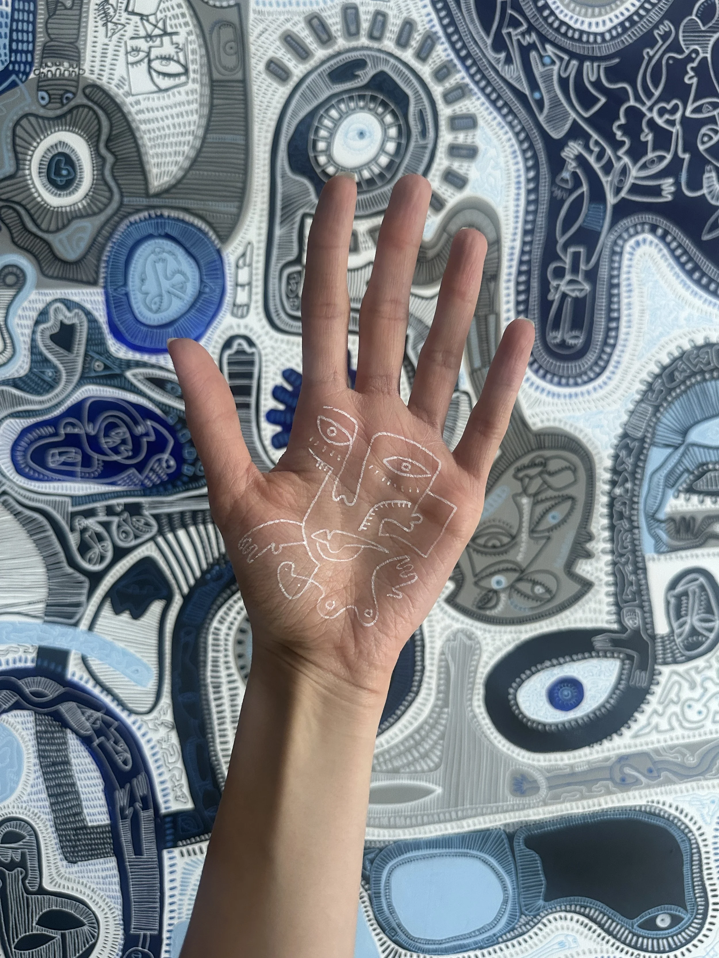 A hand with white line art tattoos of faces and abstract designs is in front of a painted wall with intricate blue, white, and black patterns including faces, eyes, and geometric shapes.