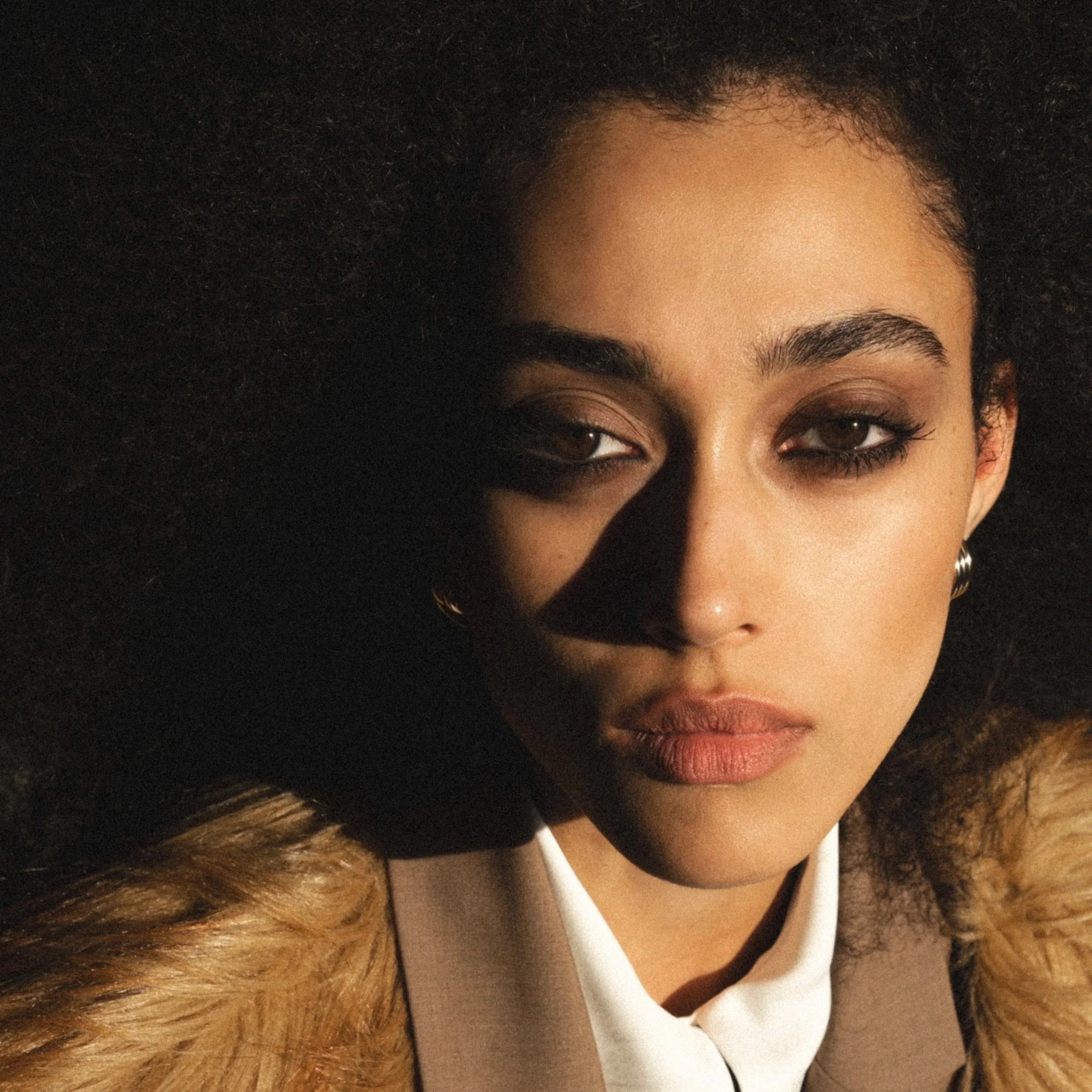 A woman with curly hair partially visible, wearing makeup, a white shirt, and hoop earrings.