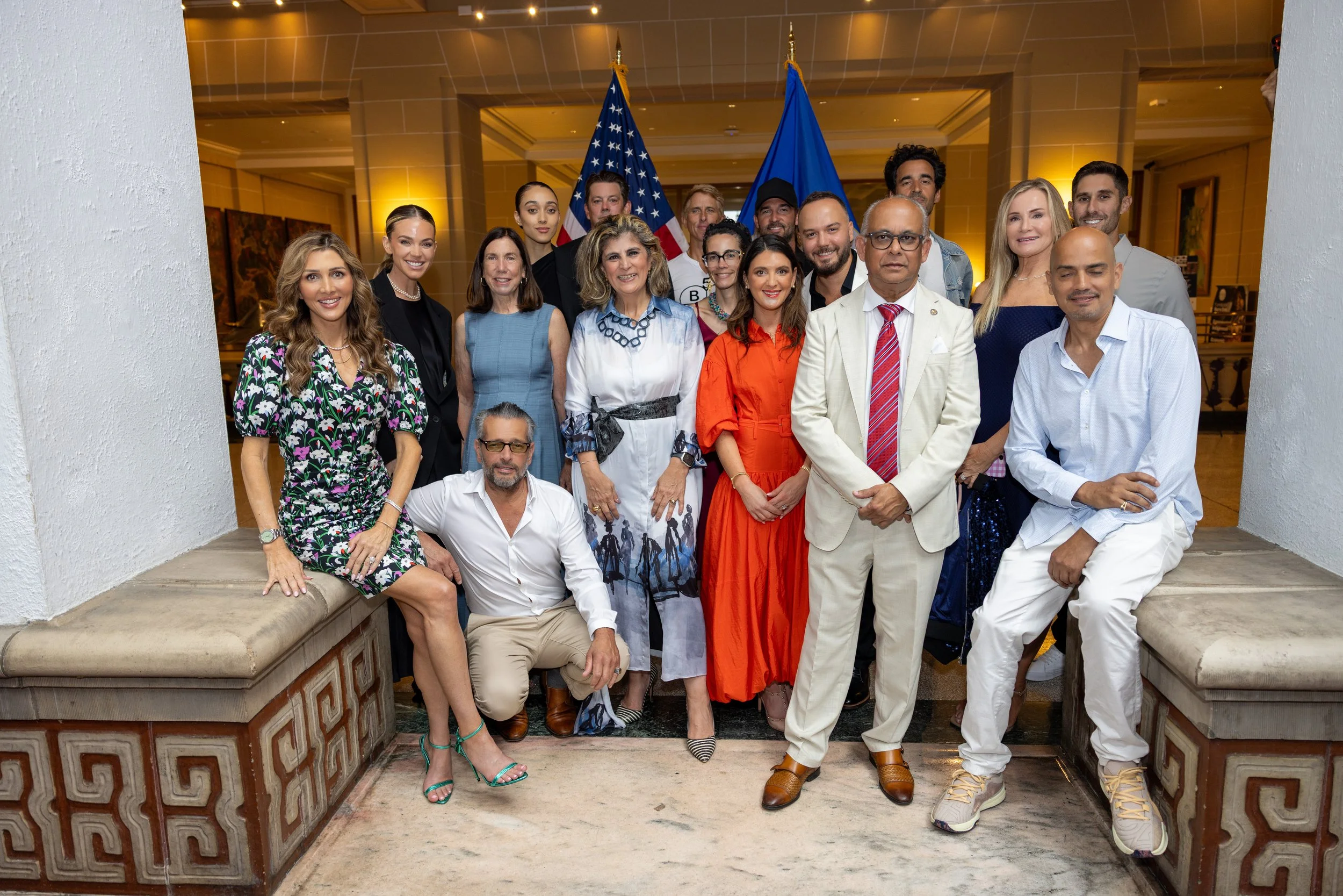 A diverse group of people posing together at an event, with two flags in the background, American and blue flag, inside a well-lit indoor space.