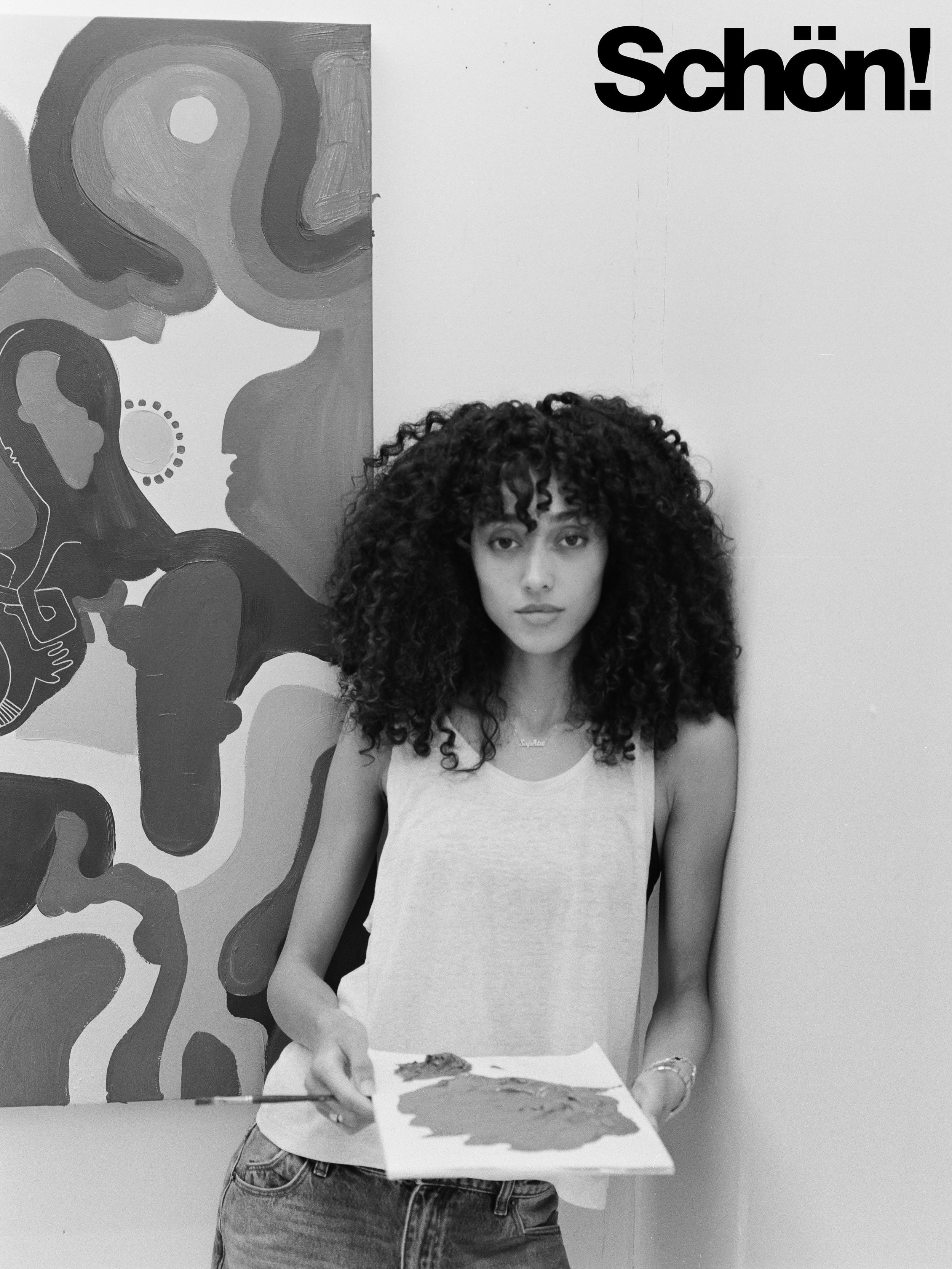 A woman with curly hair wearing a light-colored tank top and jeans, standing in front of a wall with a large abstract black and white mural, holding a piece of paper or artwork, with the word "Schön!" in large black letters on the wall.