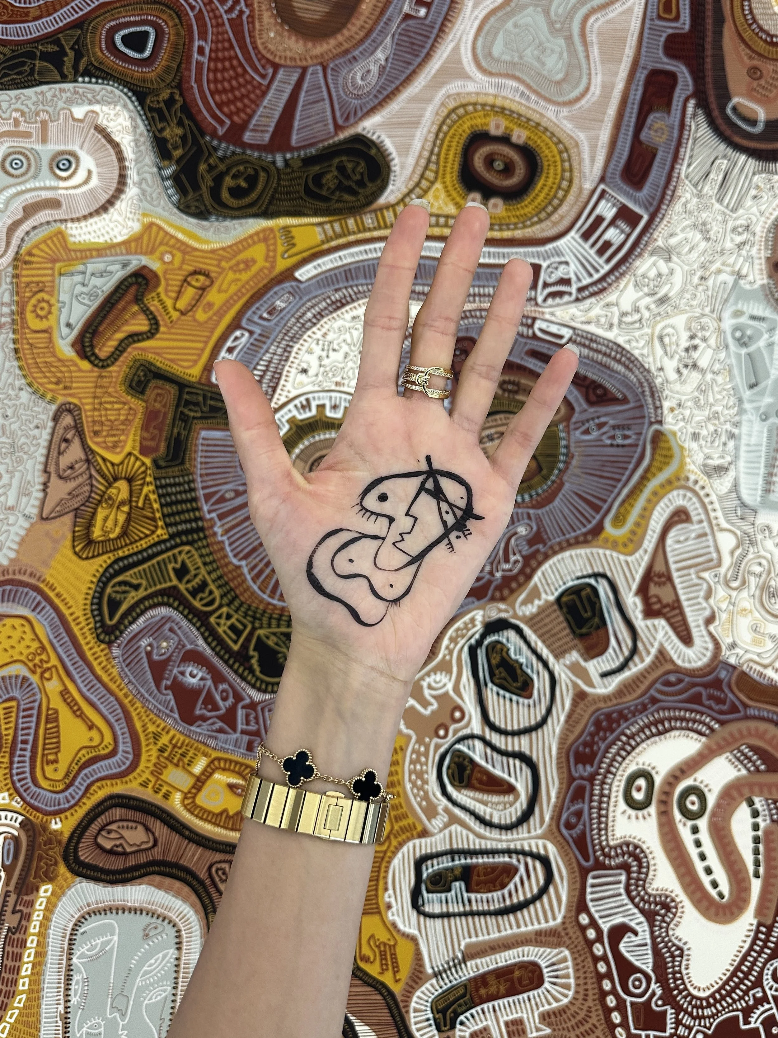 A hand with a face tattoo in black ink and three gold rings shows a peace sign in front of a colorful, abstract, patterned background. The hand is wearing a gold bracelet with black clovers.