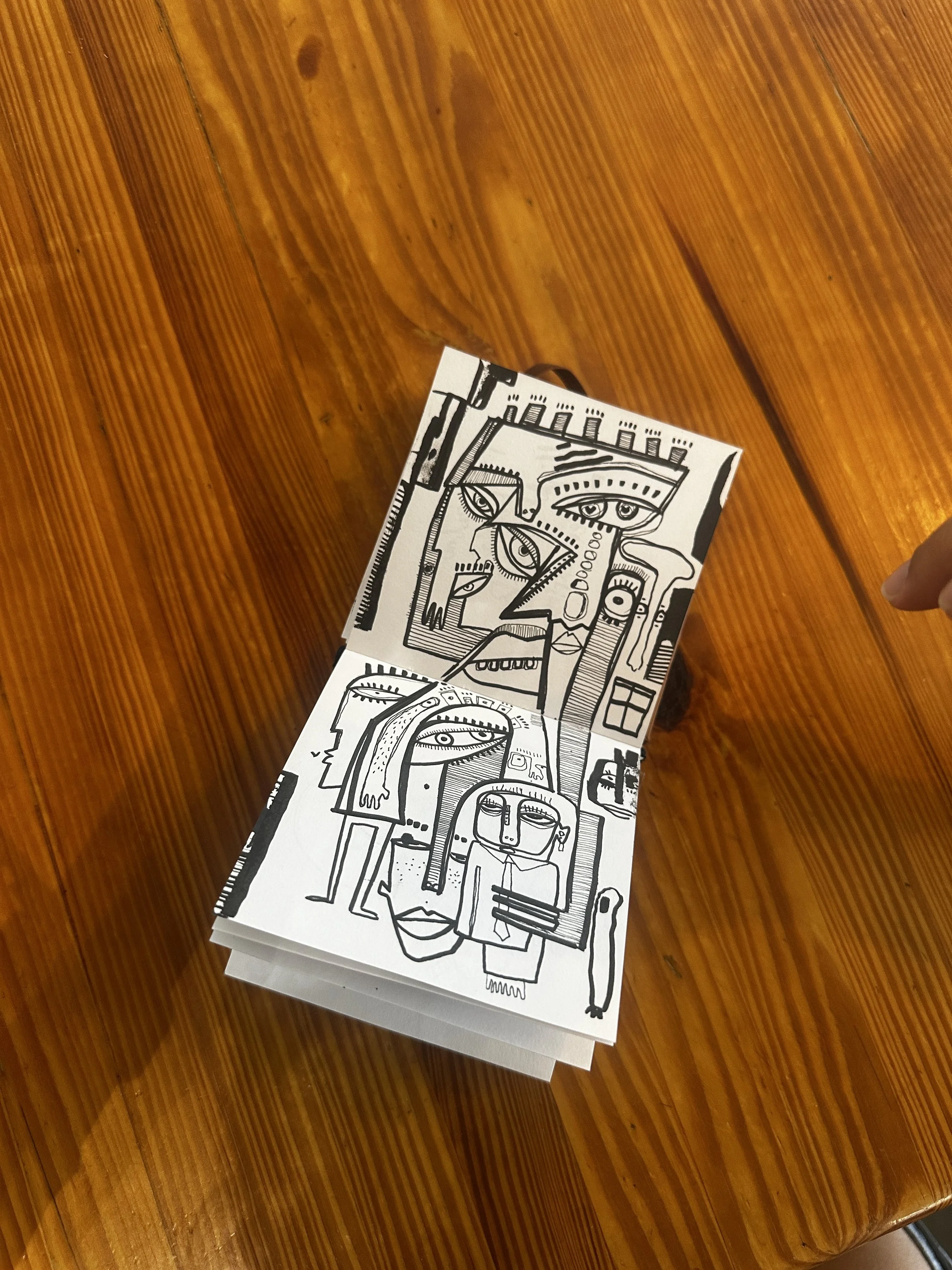 A black and white drawing with abstract human faces and figures, featuring multiple eyes and distorted features, on a stack of papers placed on a wooden surface.