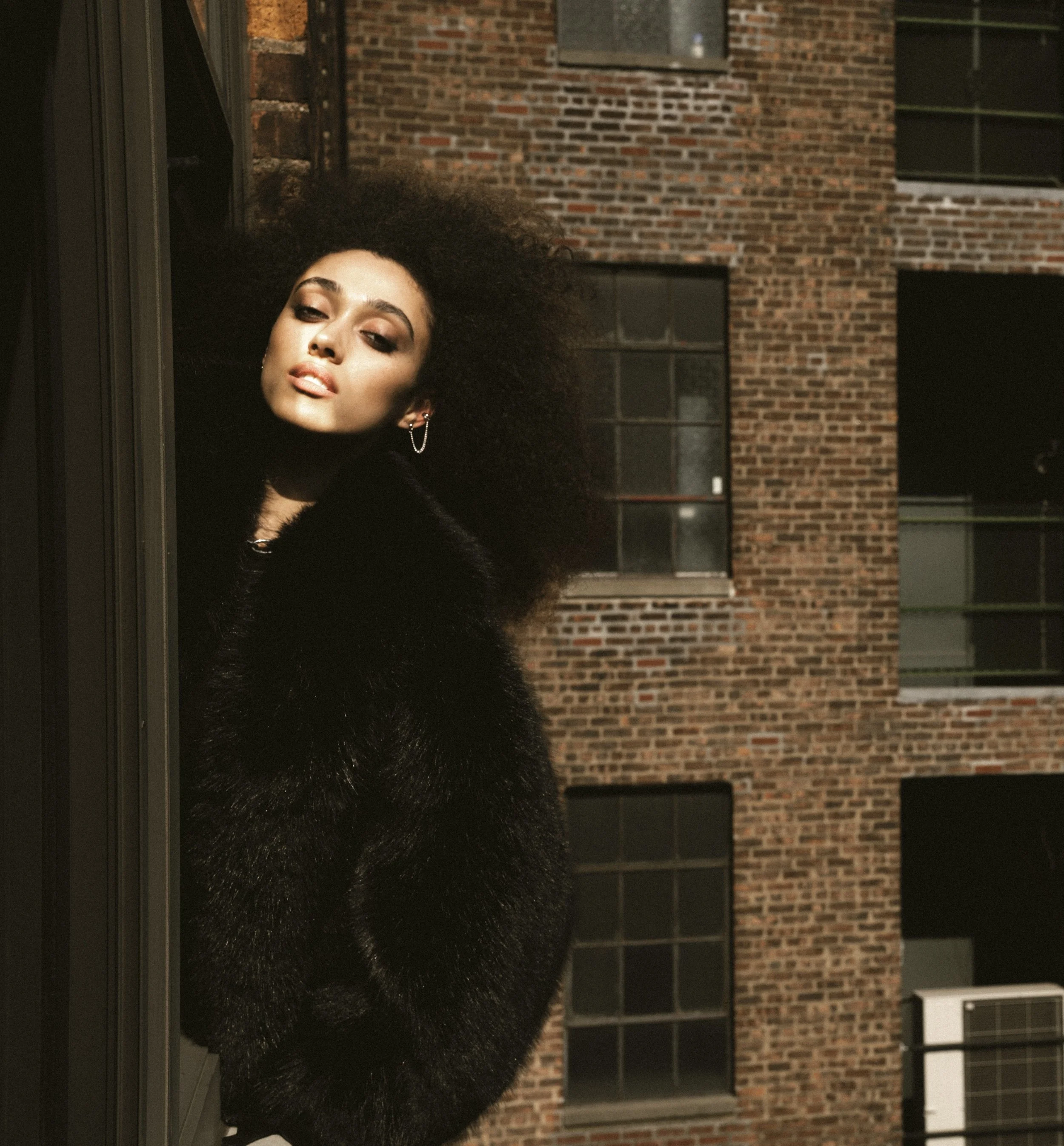 A woman with dark, curly hair and earrings leans out from behind a door on a balcony at night, with brick buildings in the background.